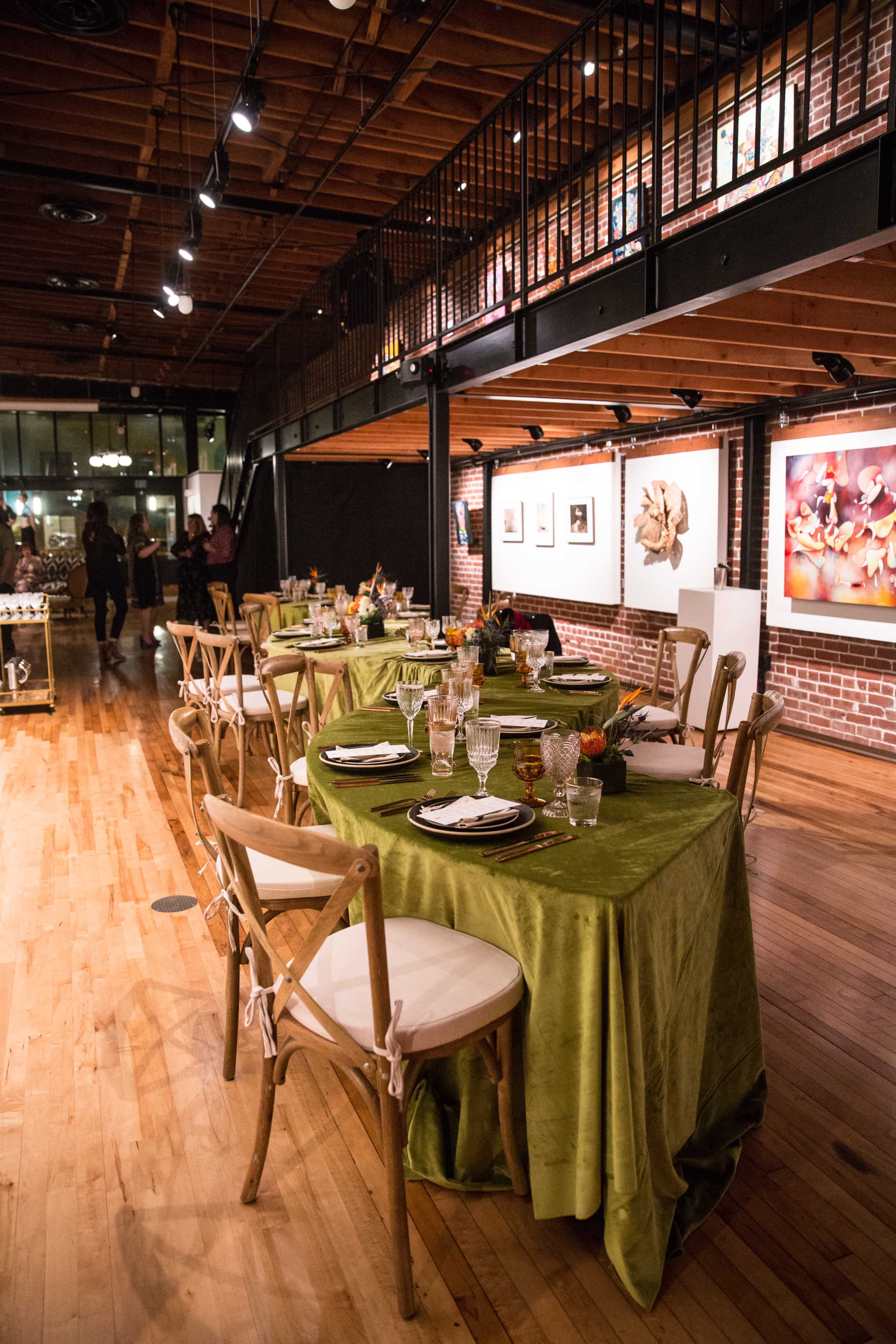 A dining space featuring wooden tables set with green tablecloths and decor, under a wooden ceiling with exposed beams and artwork displayed on the walls.