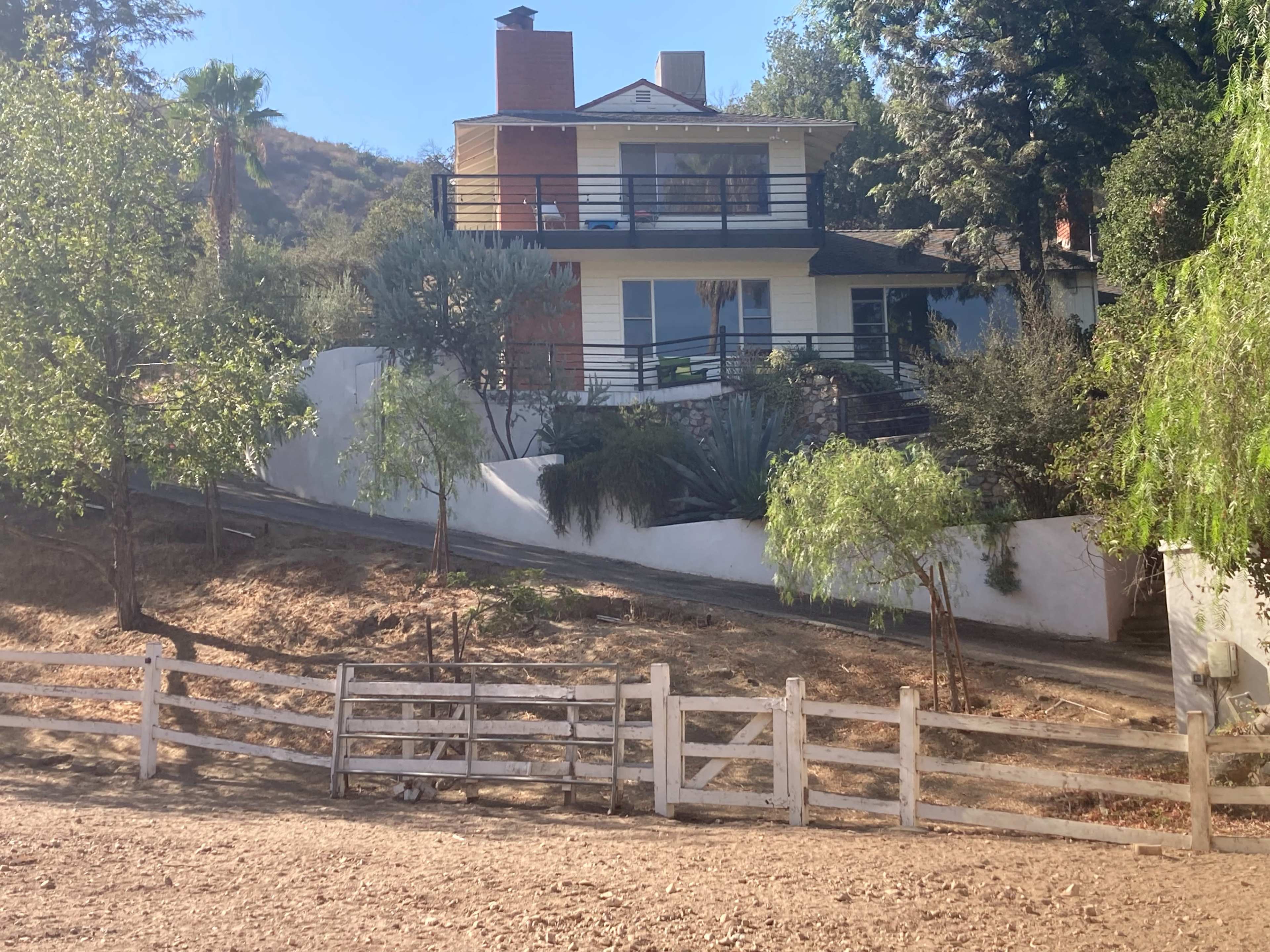 A two-story house with a balcony is situated on a hillside, surrounded by trees and a white fence.