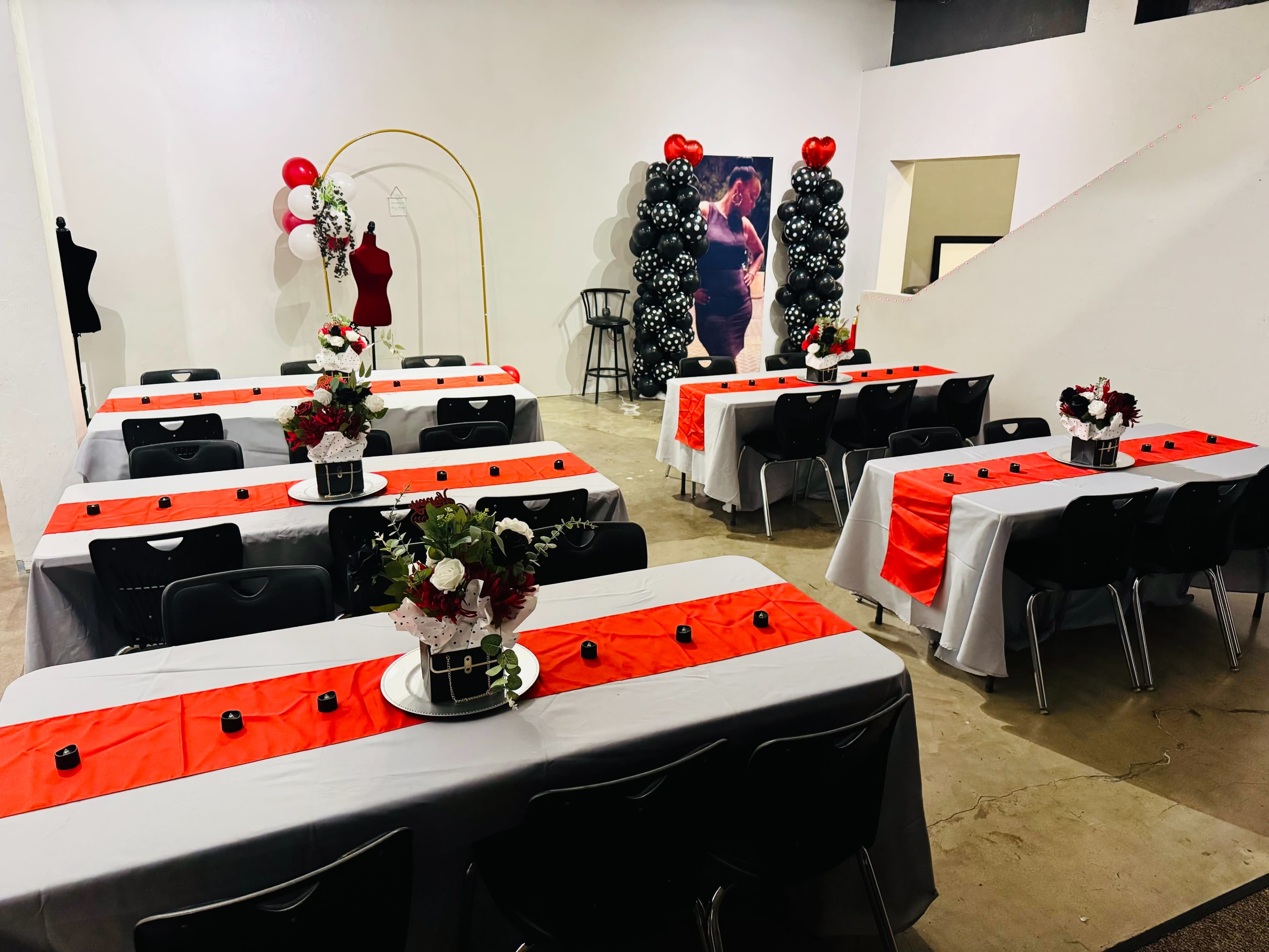 A decorated event space features several tables arranged with red table runners, floral centerpieces, and black candles, alongside balloon arches in the background.