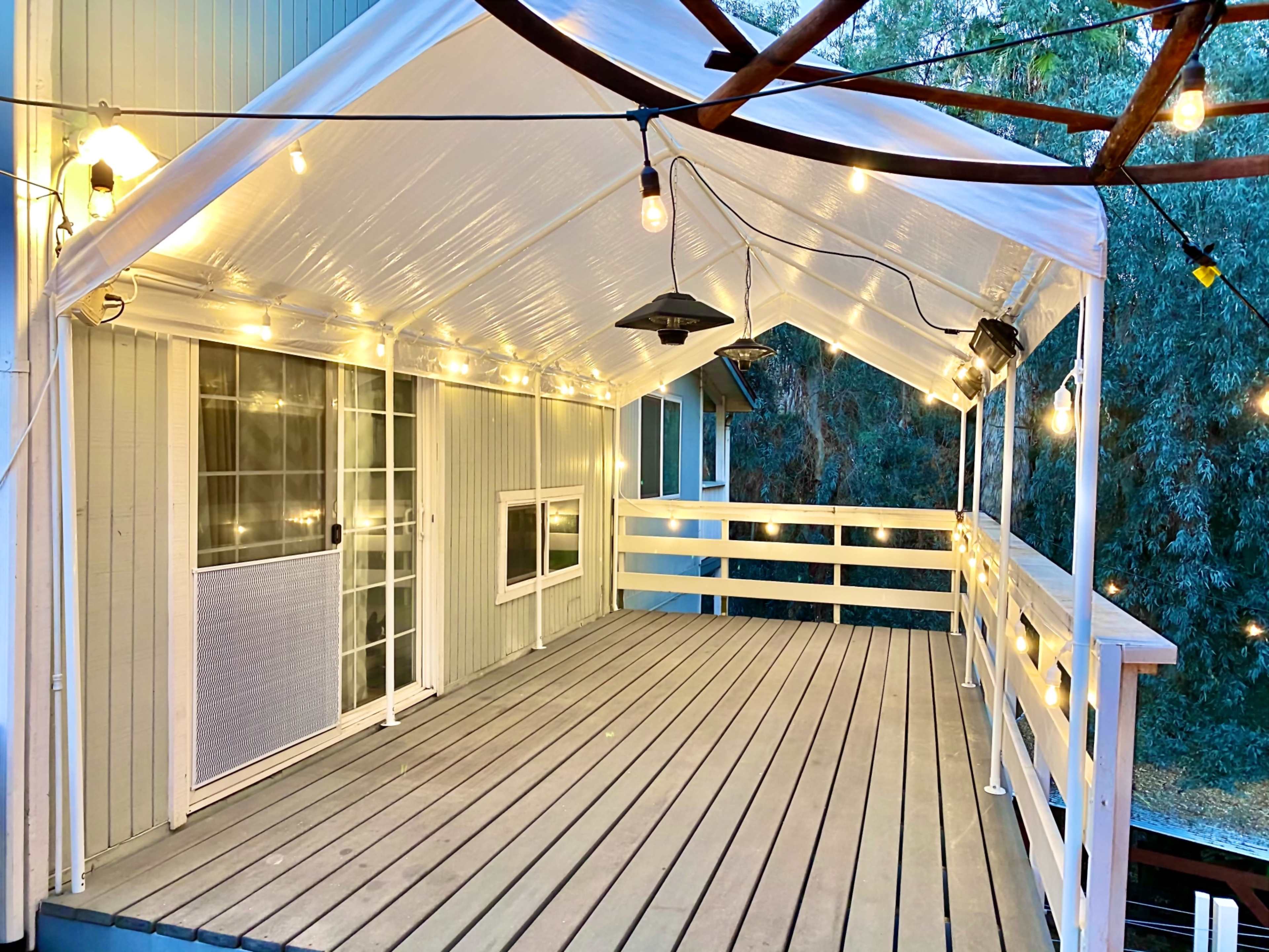 The image shows a covered outdoor deck with string lights and a wooden floor, surrounded by greenery.