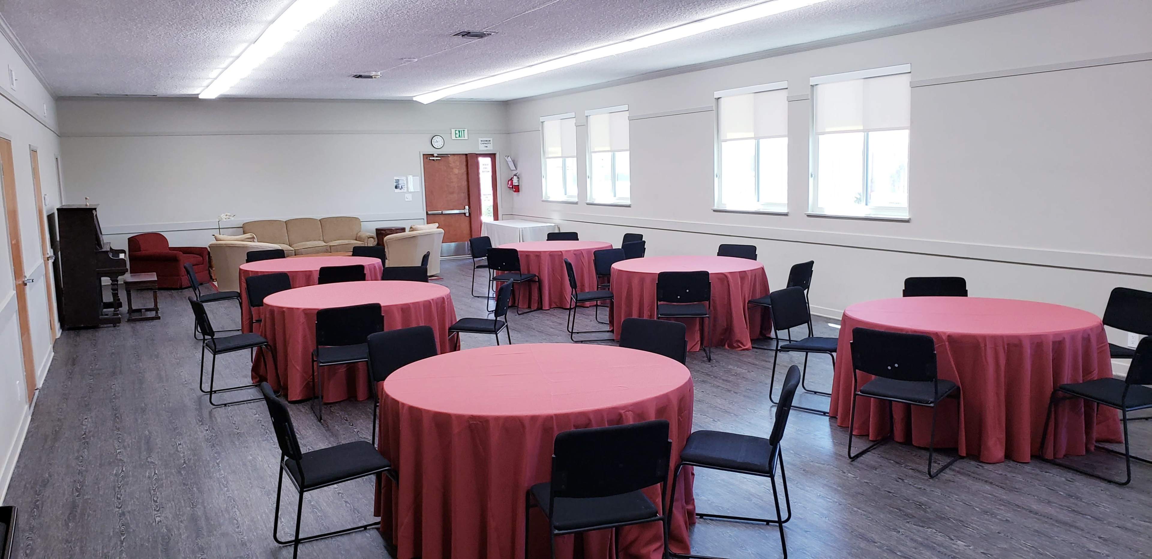 The image shows a room set up for an event, featuring several round tables covered with red tablecloths and surrounded by black chairs.