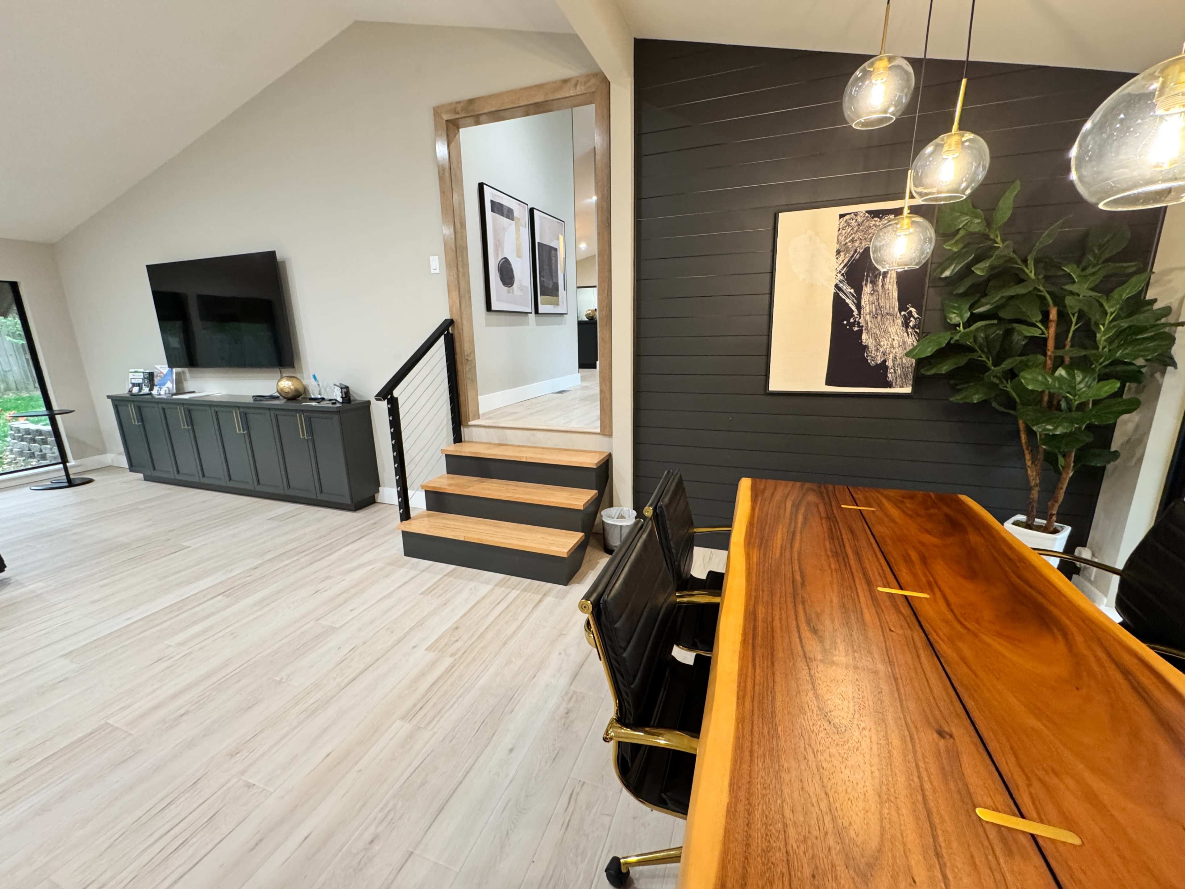 The image shows a modern dining area featuring a wooden table with black chairs, a staircase leading to another room, and a large plant beside the wall.