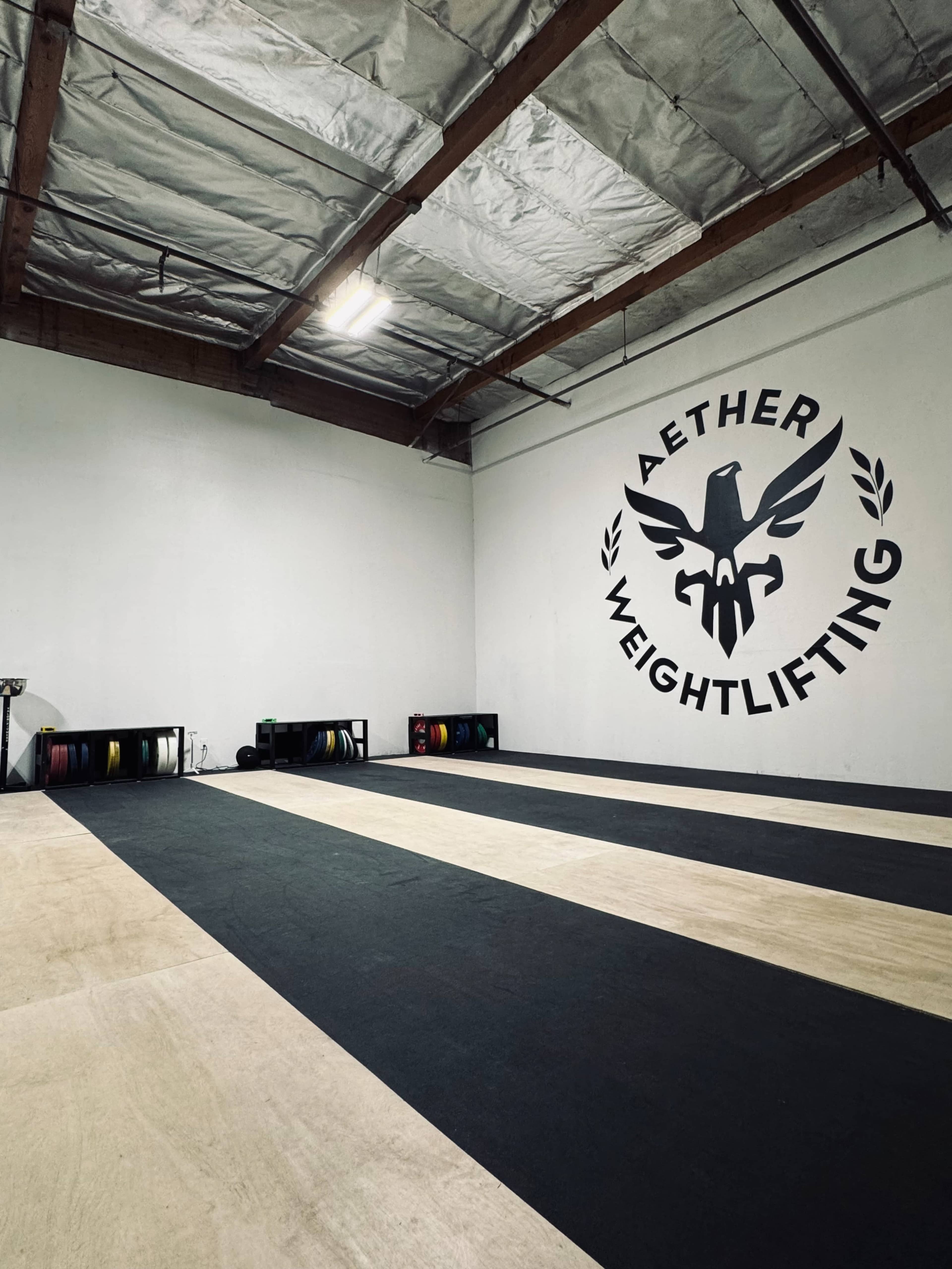 The image shows an indoor gym space with a large wall logo for "Aether Weightlifting," featuring a stylized eagle, and equipped with weightlifting platforms and equipment.