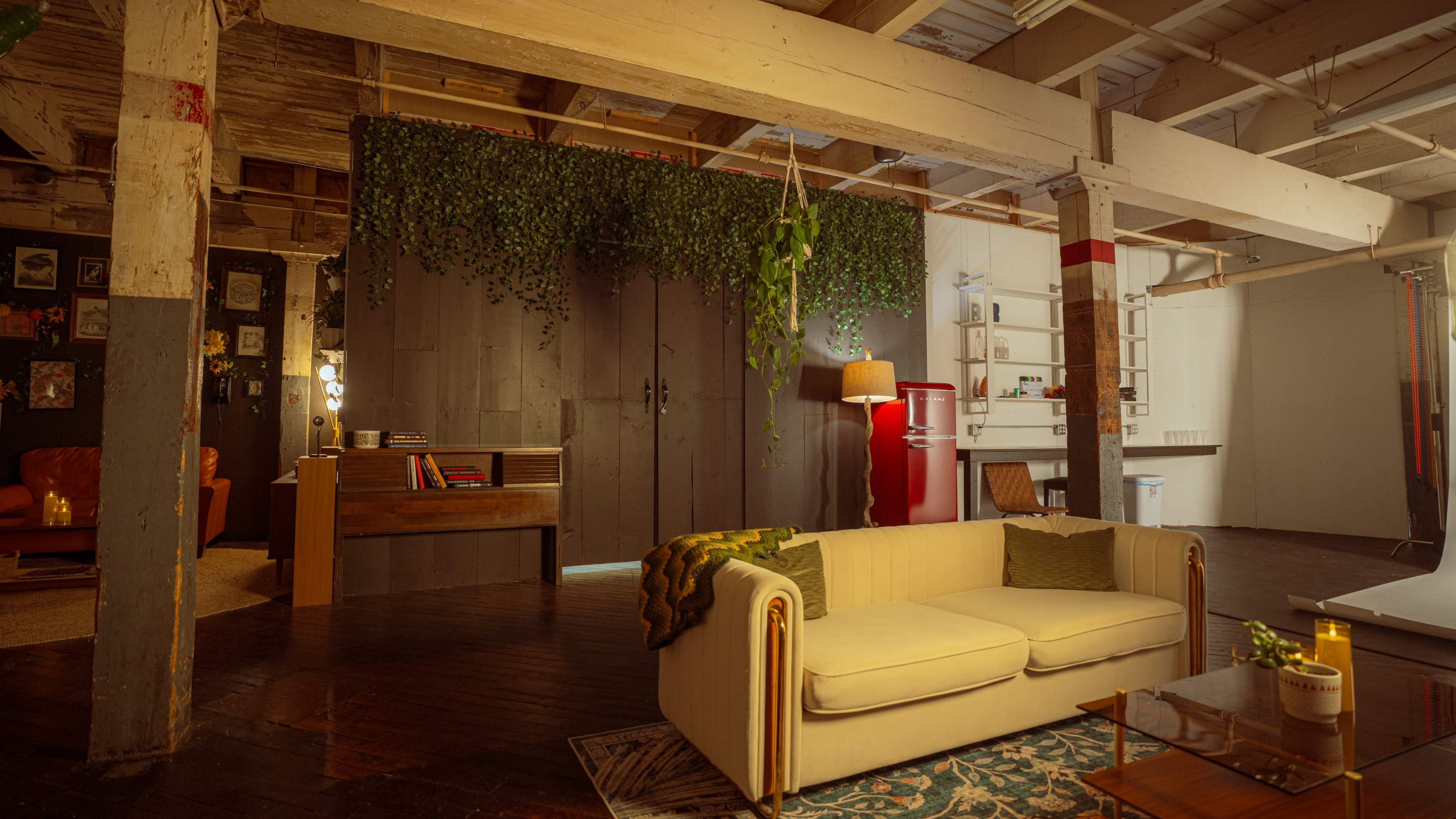 The image features a spacious industrial-style living area with a white couch, a wooden divider adorned with greenery, and a retro red refrigerator in the background.