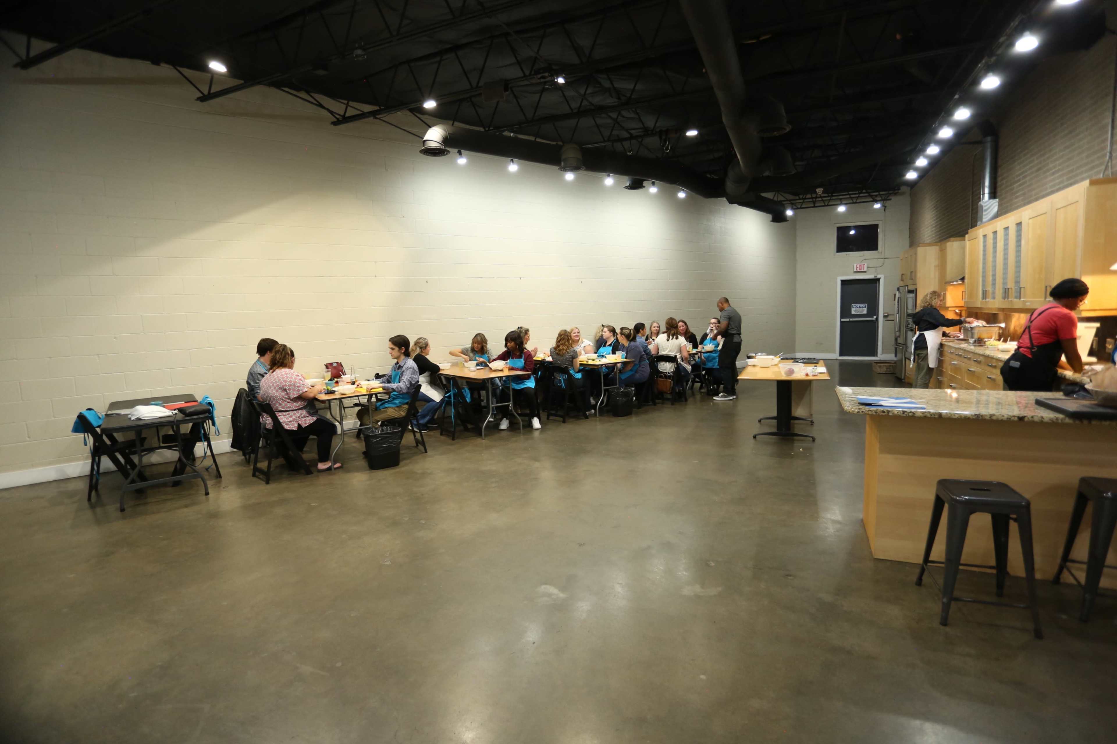 A group of people sits at tables in a well-lit room with a kitchen area in the background.
