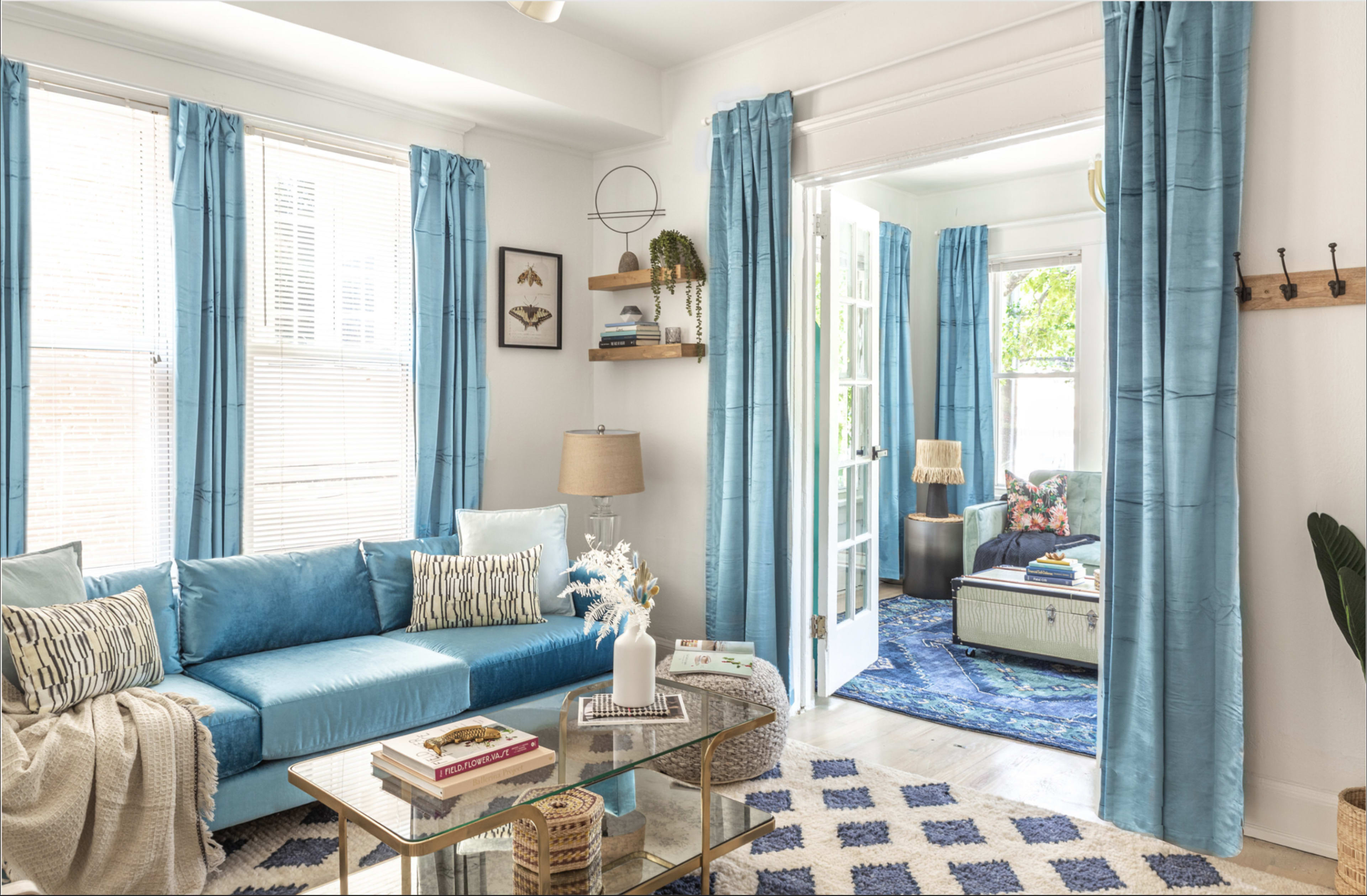 A living room features a blue sofa, light blue curtains, a glass coffee table, and a doorway leading to another room with floral decor.