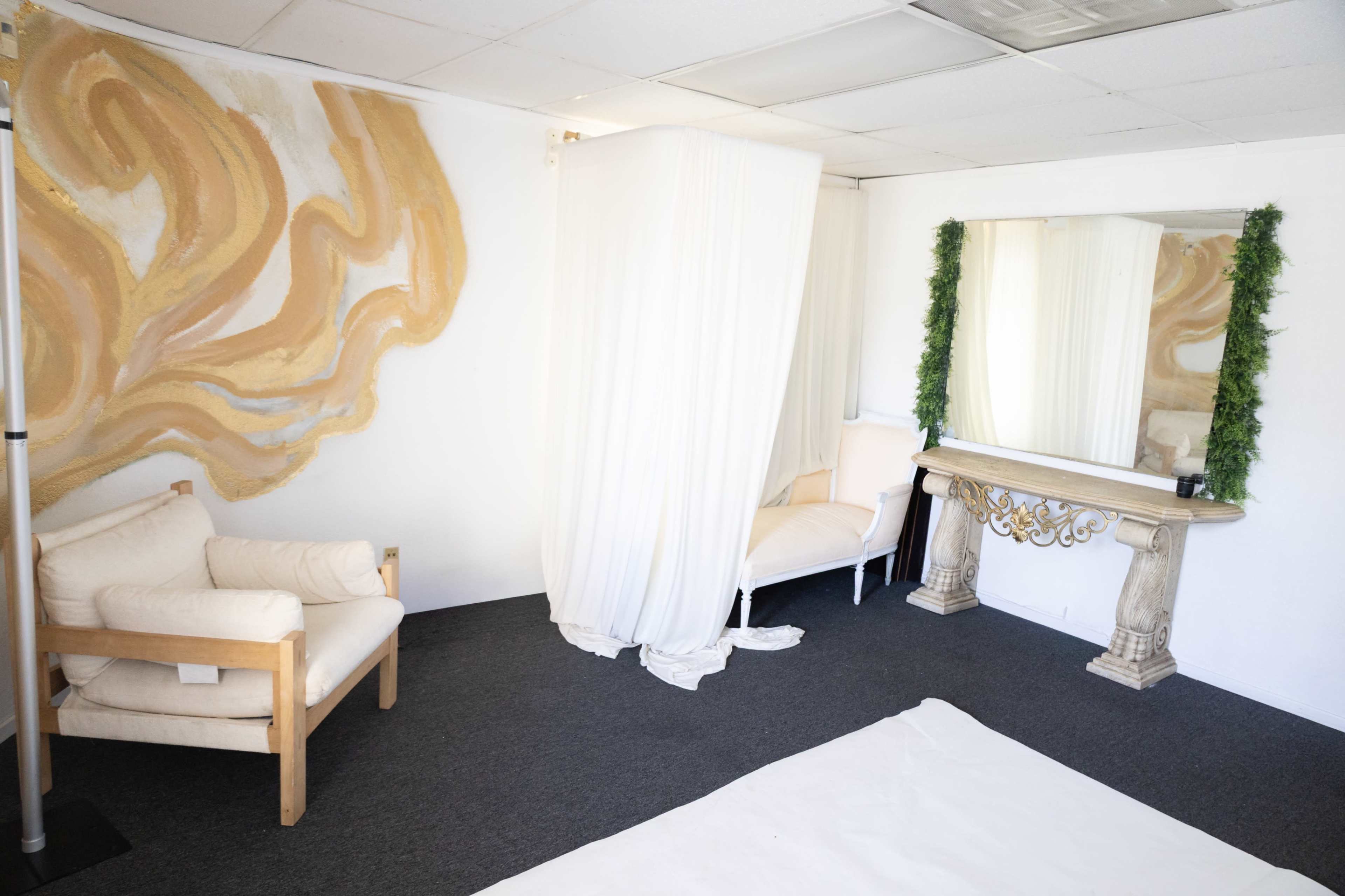 The image shows a room with a mural on one wall, two seating areas with white fabric drapes, and a decorative mirror above a stone-like console table.