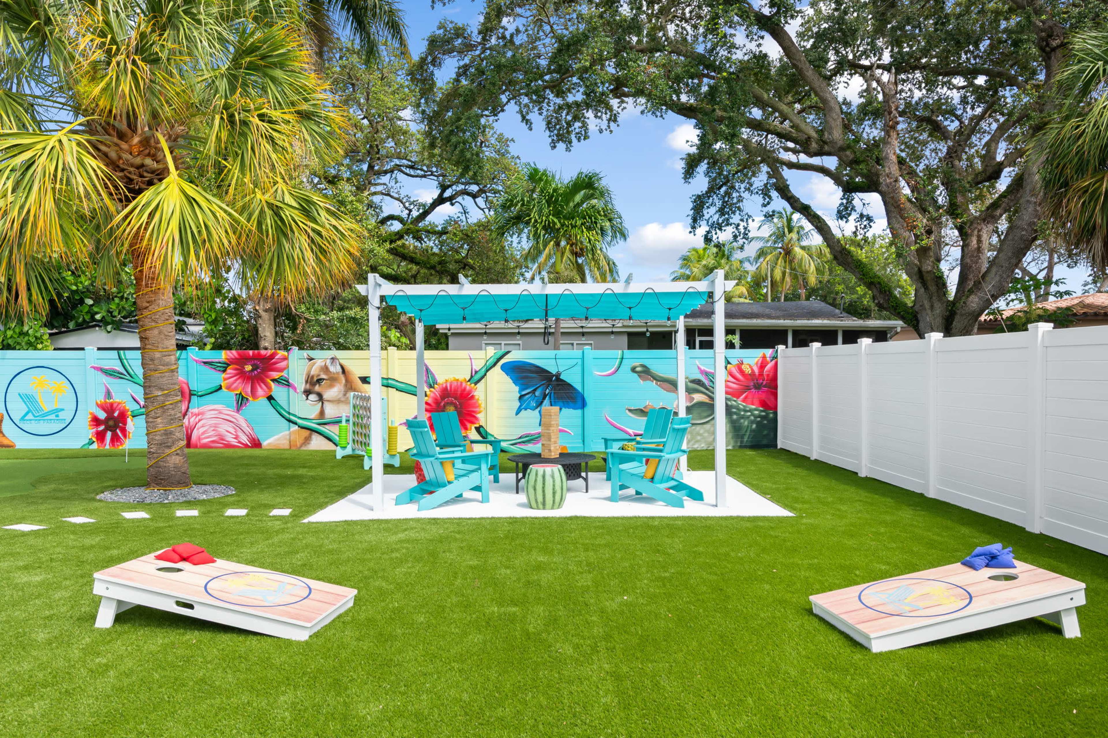 A colorful outdoor space features a white pergola with teal chairs, a vibrant mural on the fence, and two cornhole boards on the artificial grass.