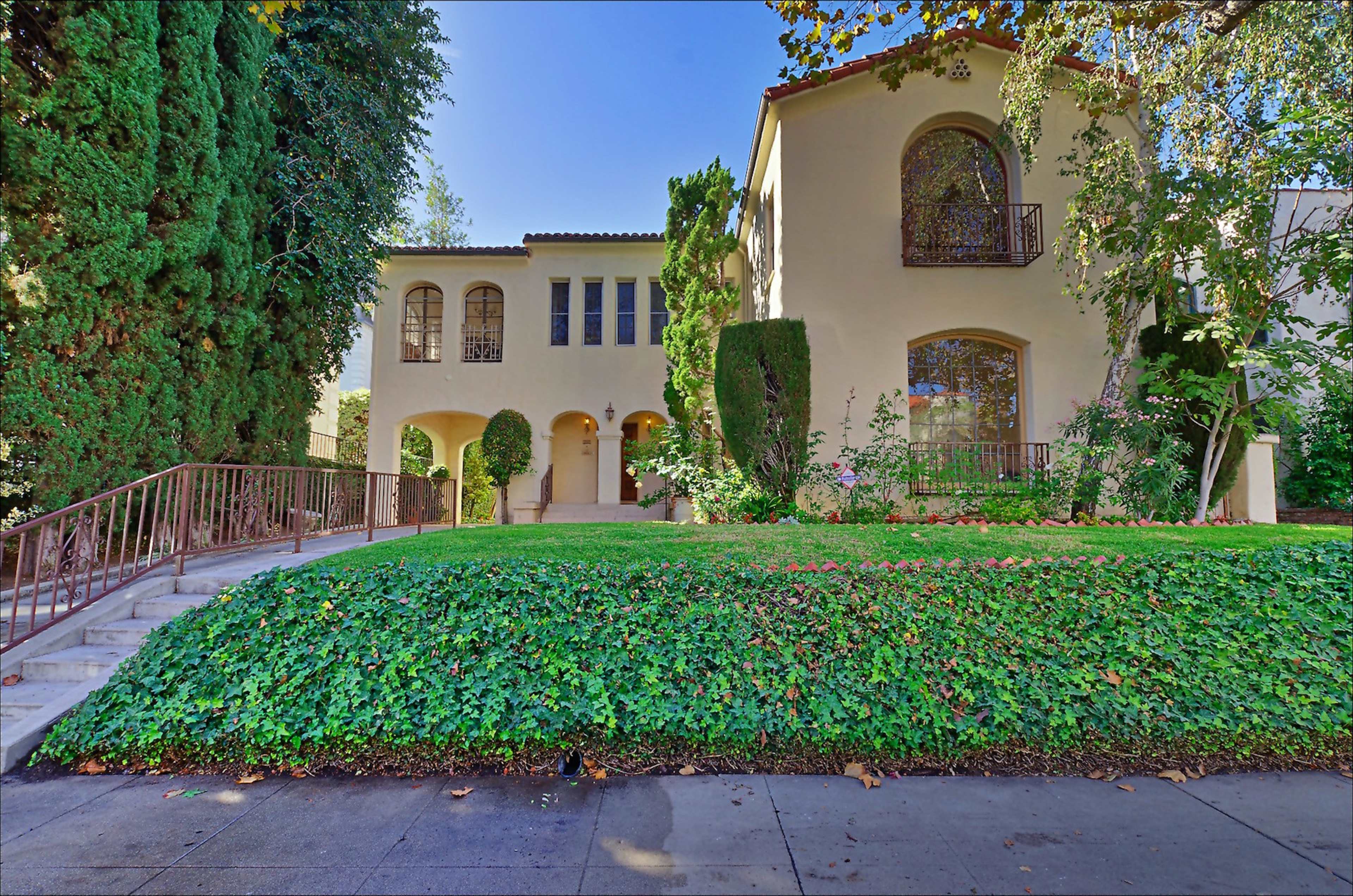 The image shows a two-story house with arched windows, surrounded by well-maintained greenery and a staircase leading to the entrance.