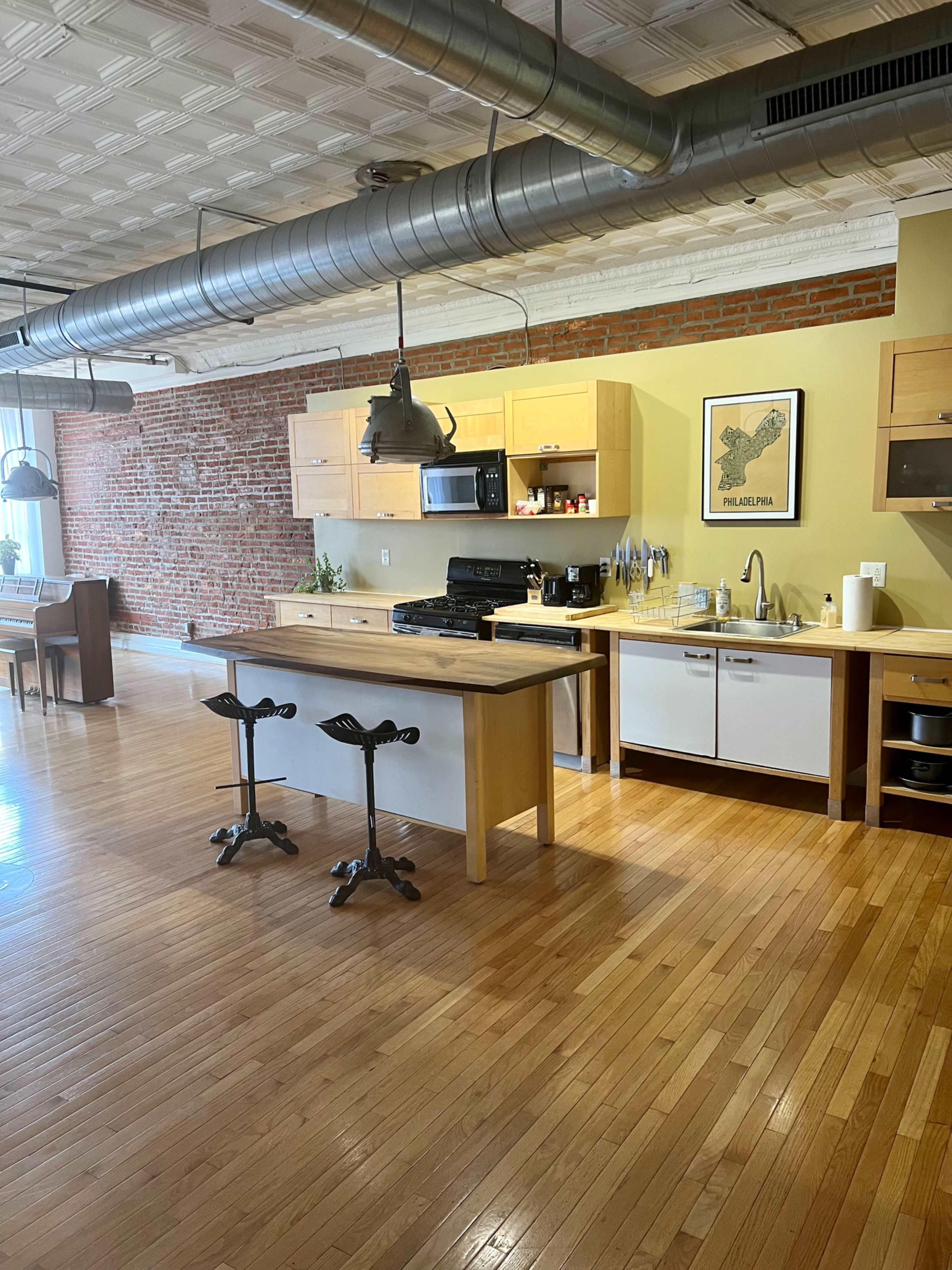 A modern kitchen with wooden cabinets, a central island, and industrial-style lighting situated in an open space with exposed brick walls and hardwood flooring.