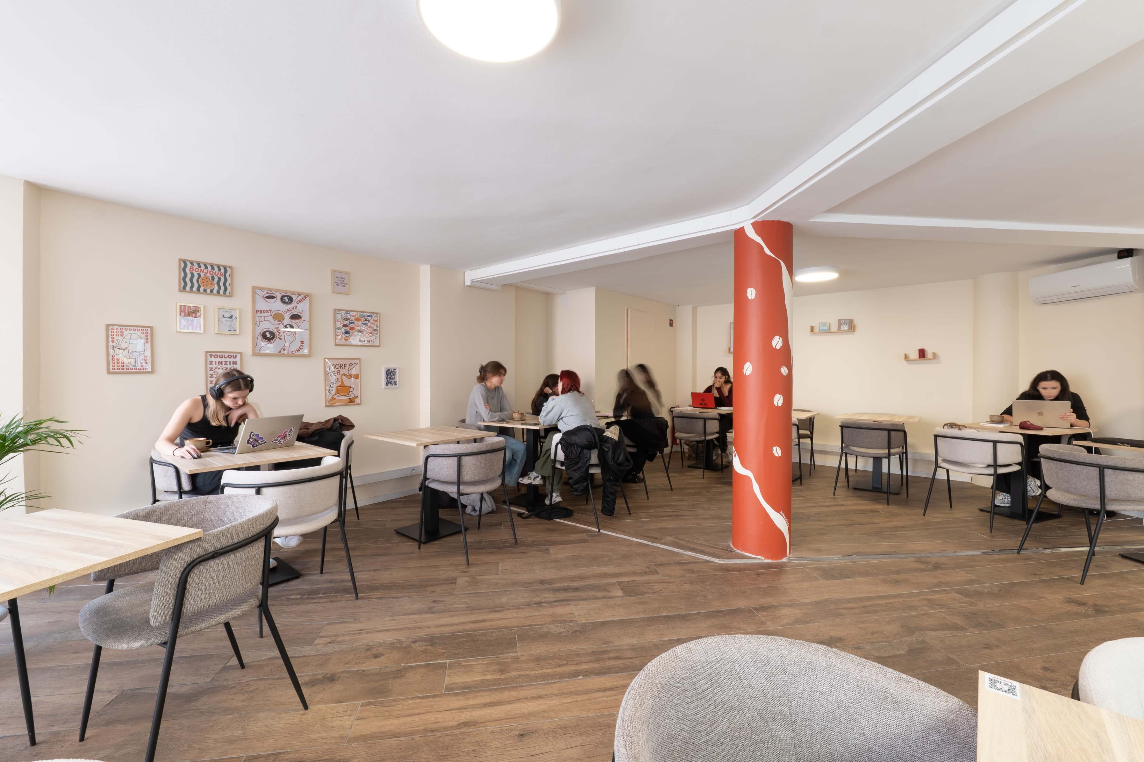 The image shows a modern coworking space with several individuals seated at tables, some engaged with laptops, and decorative artwork on the walls.