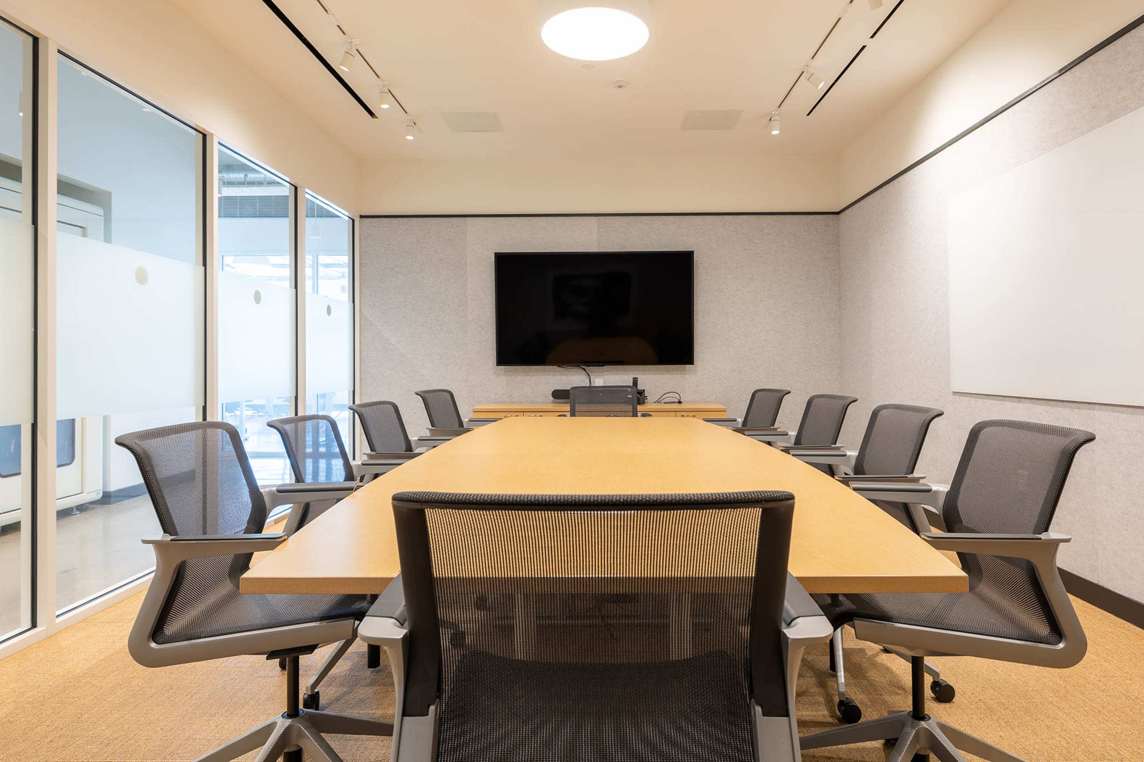 A large conference room features a long wooden table surrounded by several ergonomic chairs, with a screen mounted on the wall and large windows allowing natural light.
