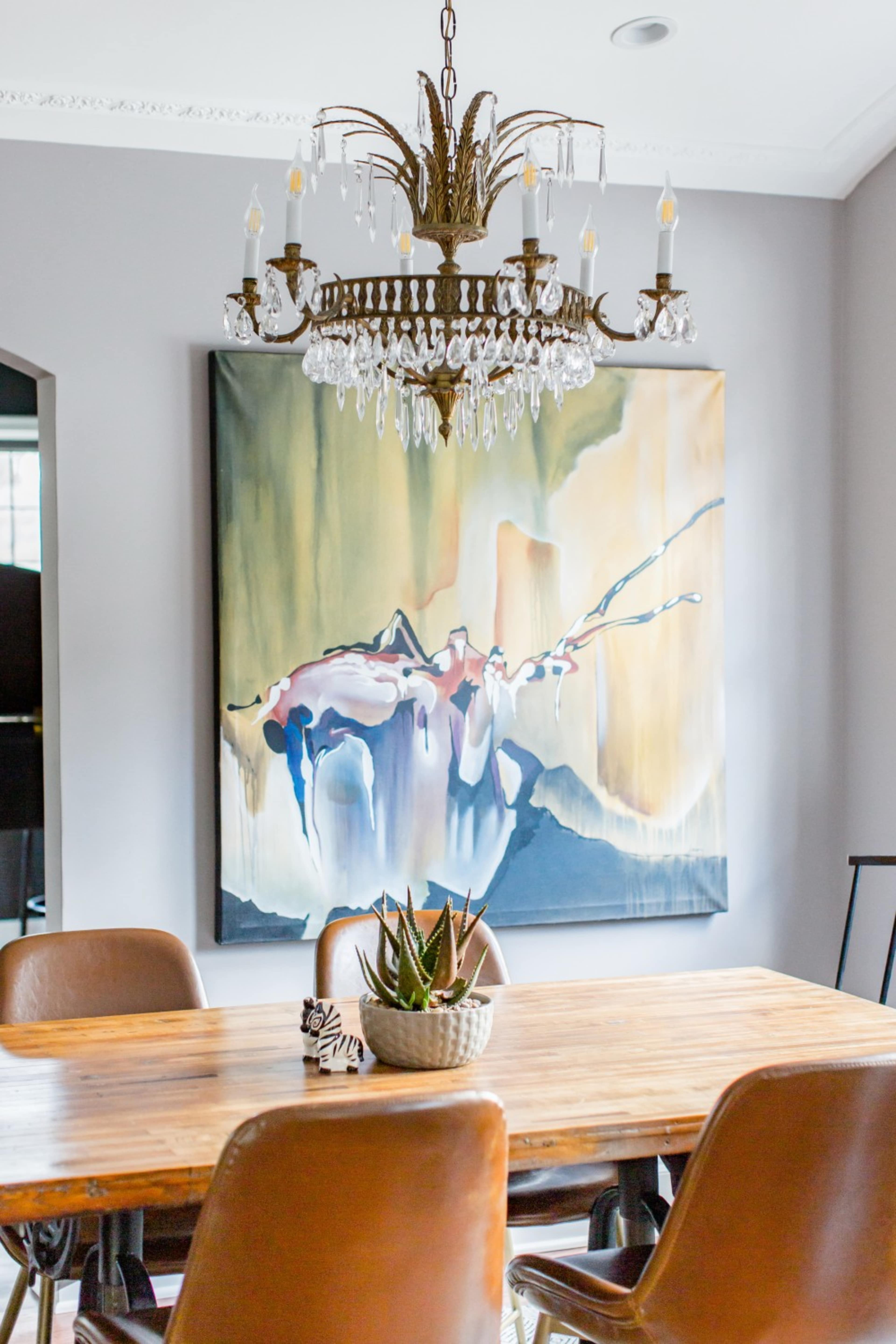 A wooden dining table is centered under a crystal chandelier with a large abstract painting hanging on the wall behind it.