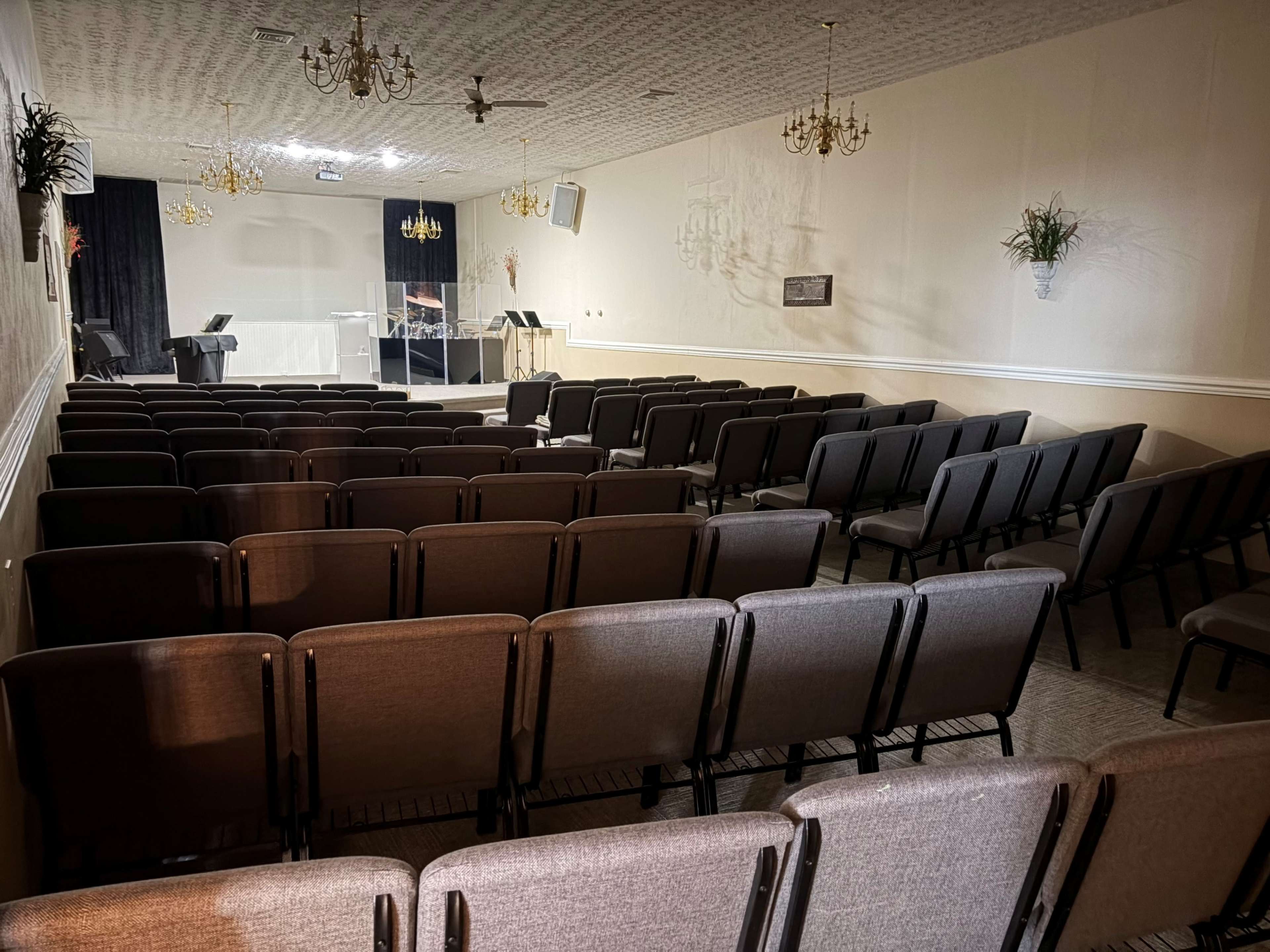 The image shows a spacious empty hall set up with rows of gray chairs, a podium, and a piano in front, illuminated by soft overhead lighting.
