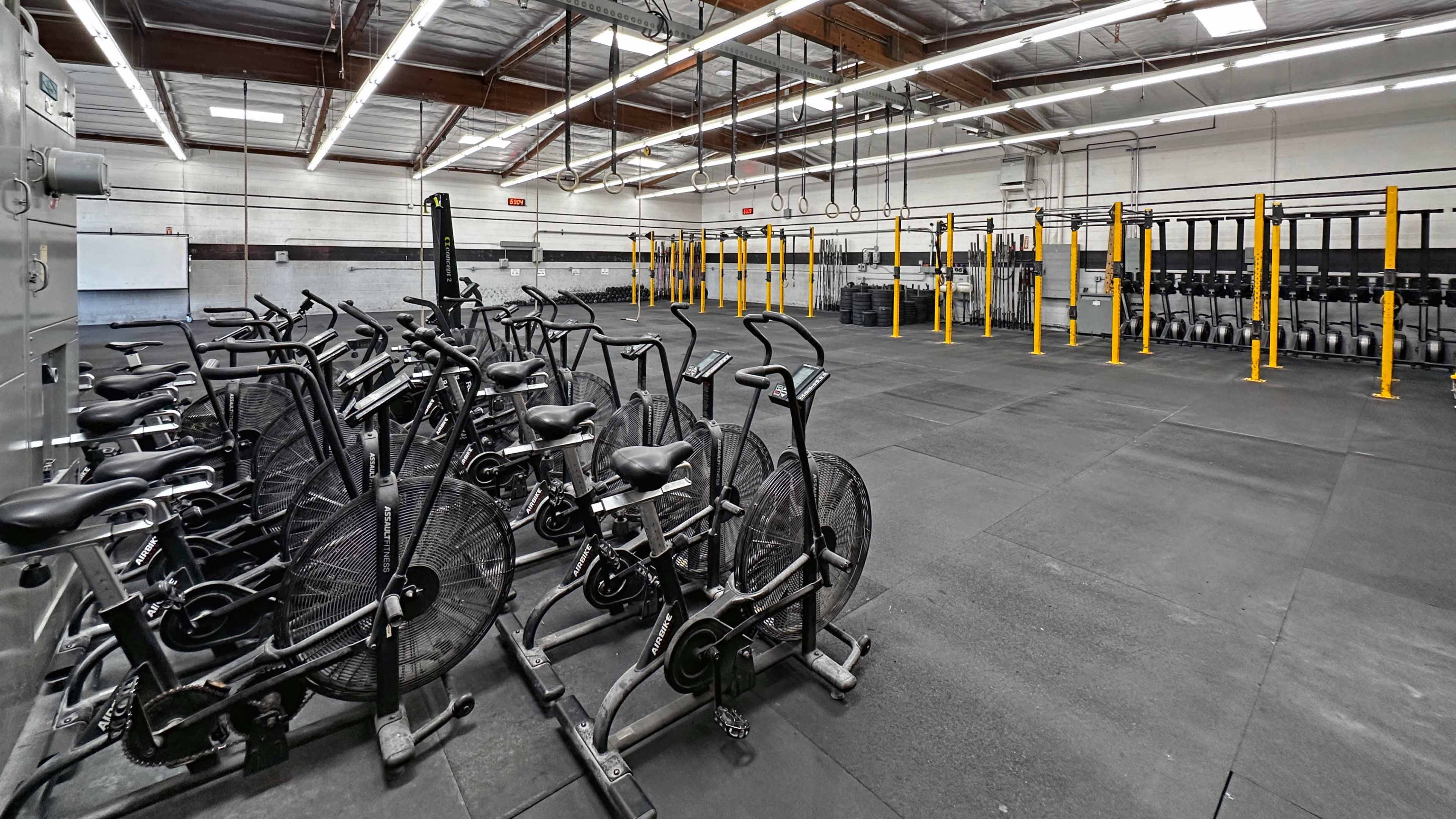 The image shows a spacious gym interior with several stationary bikes lined up on one side, and a set of yellow pull-up bars and weight equipment on the opposite side.