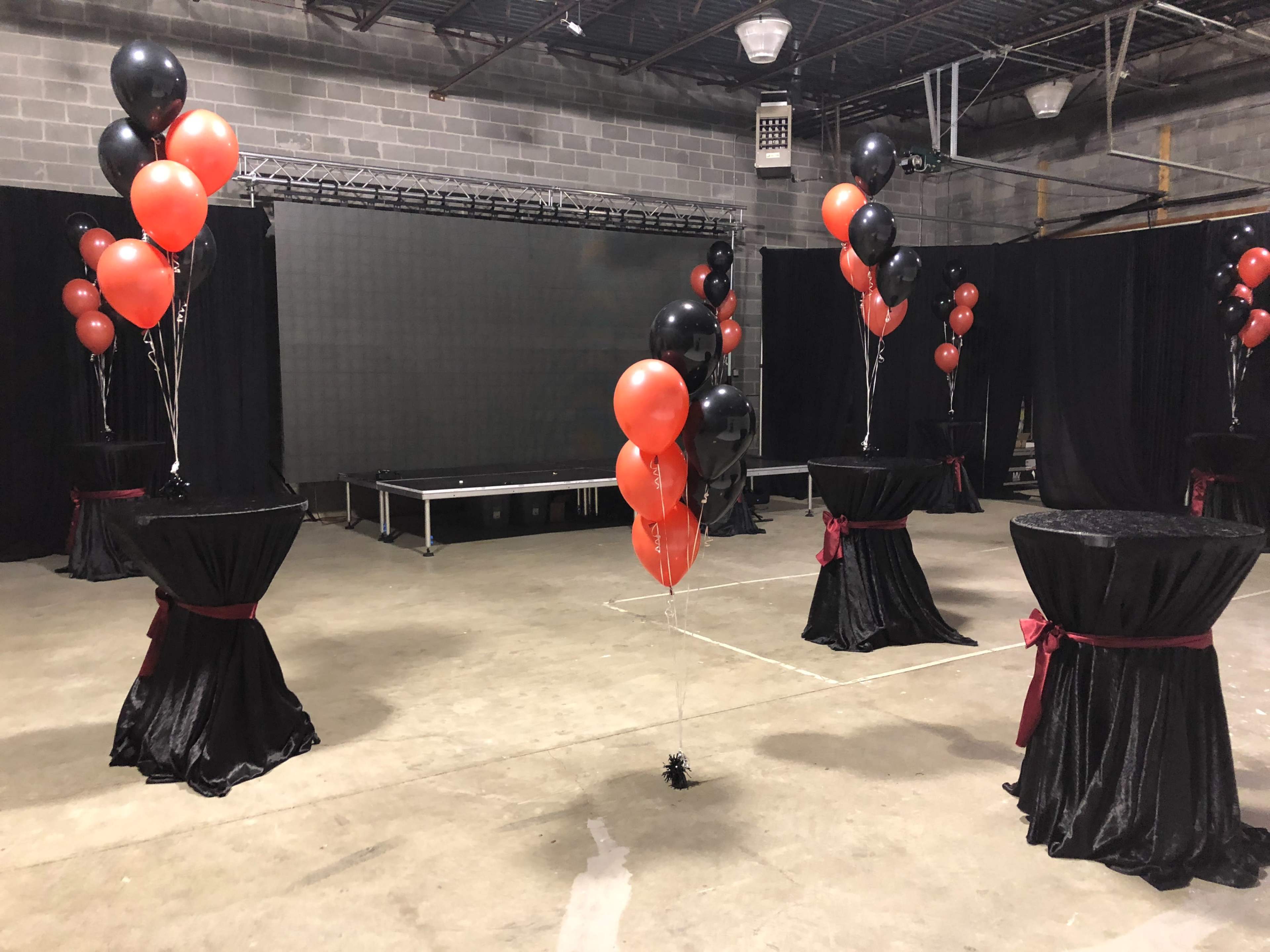 A decorated venue with black and red balloons on tables, a stage in the background, and black curtains.