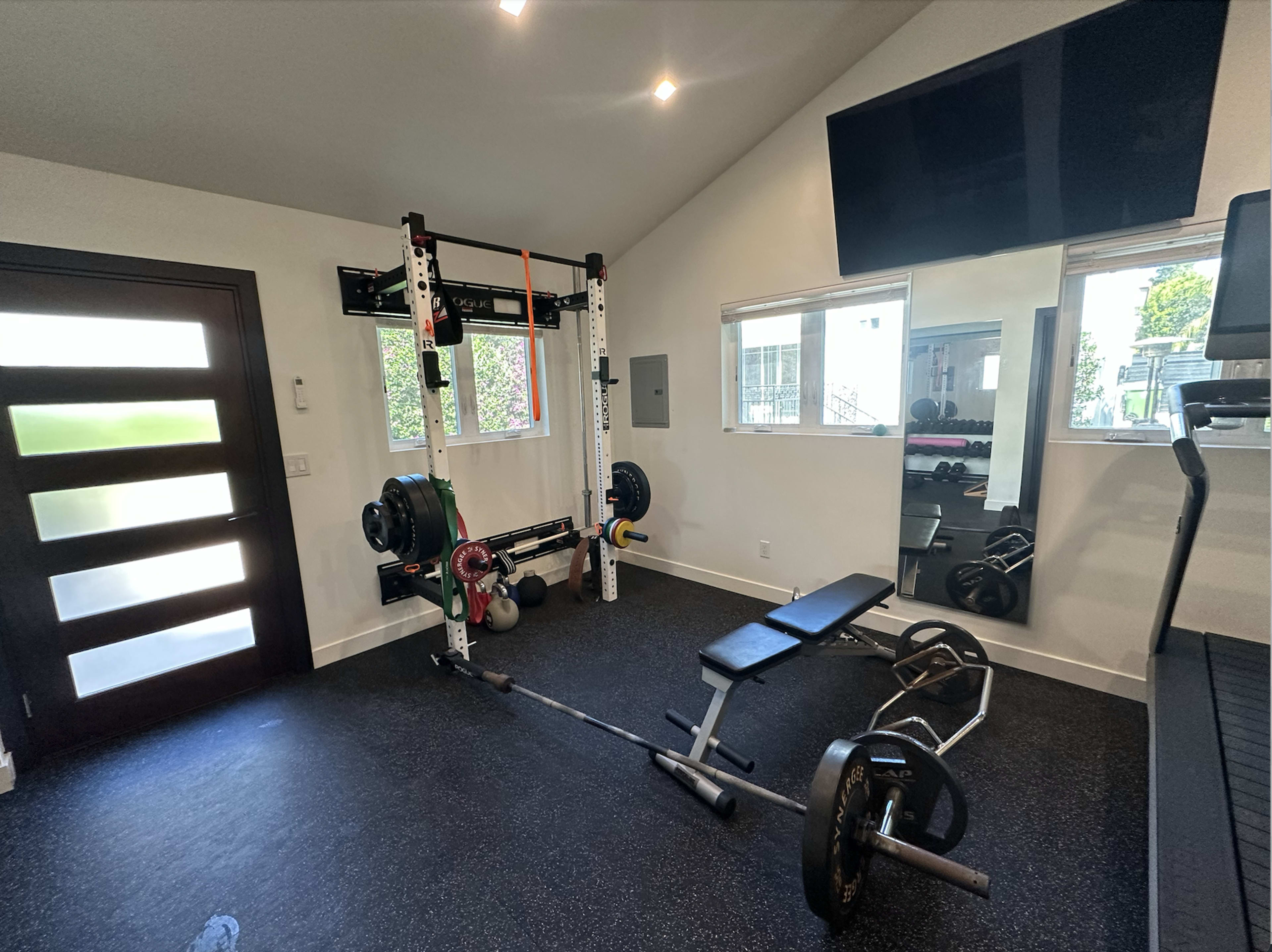 The image shows a home gym equipped with a weightlifting rack, a bench, various weights, and exercise equipment, set against a modern interior with large windows.