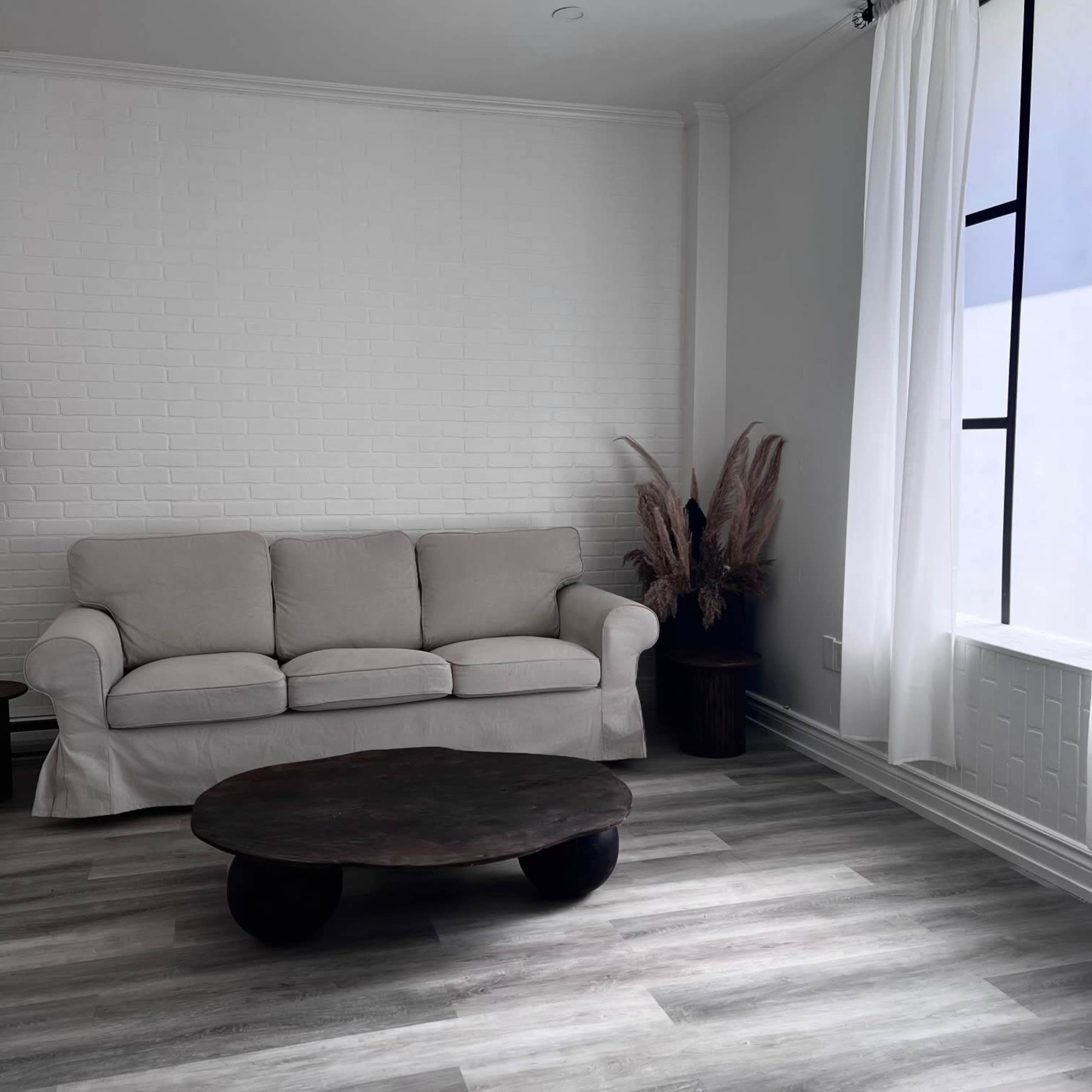 A simple living room with a light-colored sofa, a round wooden coffee table, and a window adorned with sheer curtains.