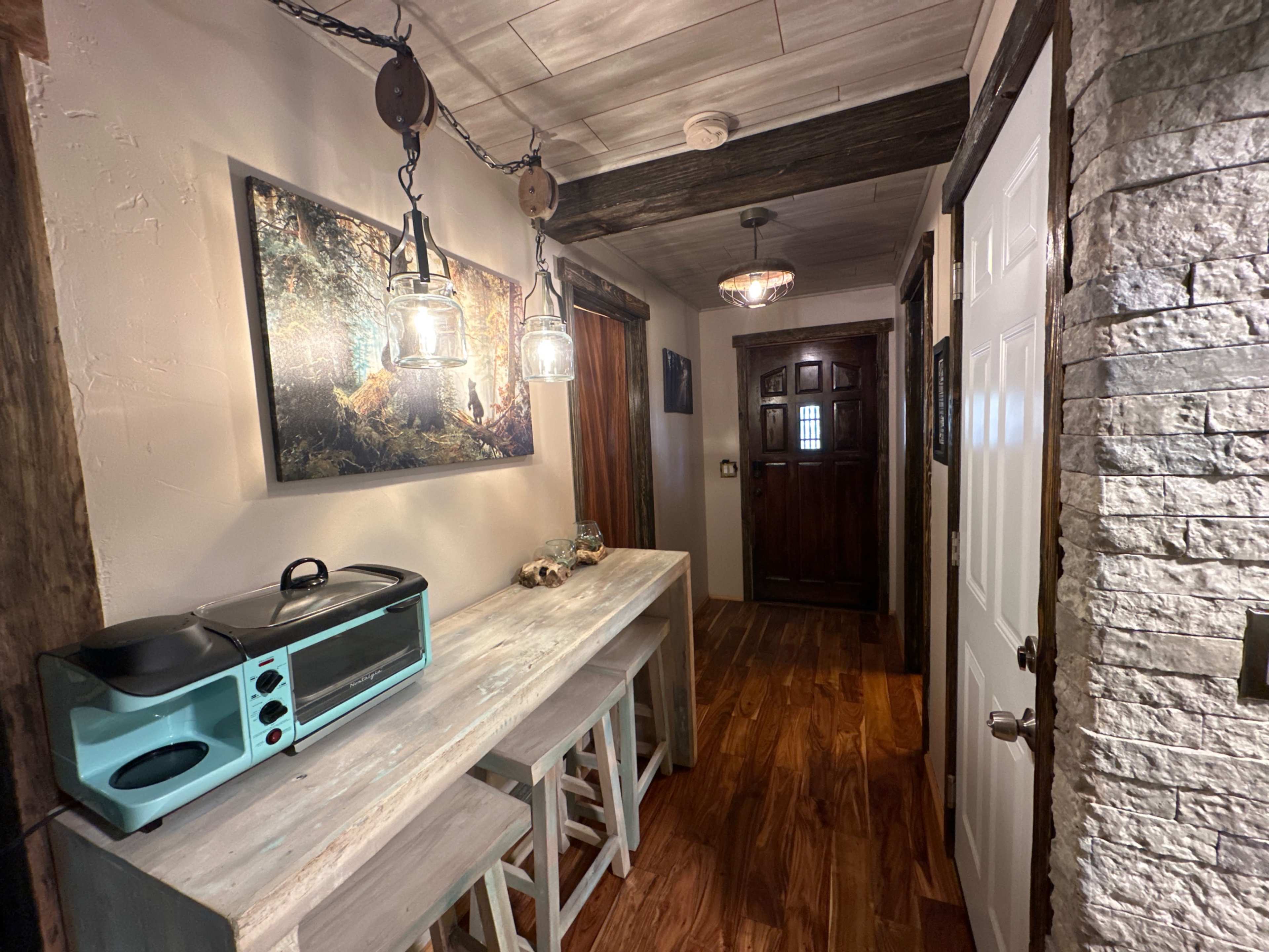 A narrow hallway with wooden flooring, a light-colored table with stools, and various light fixtures, leading to a wooden door at the end.