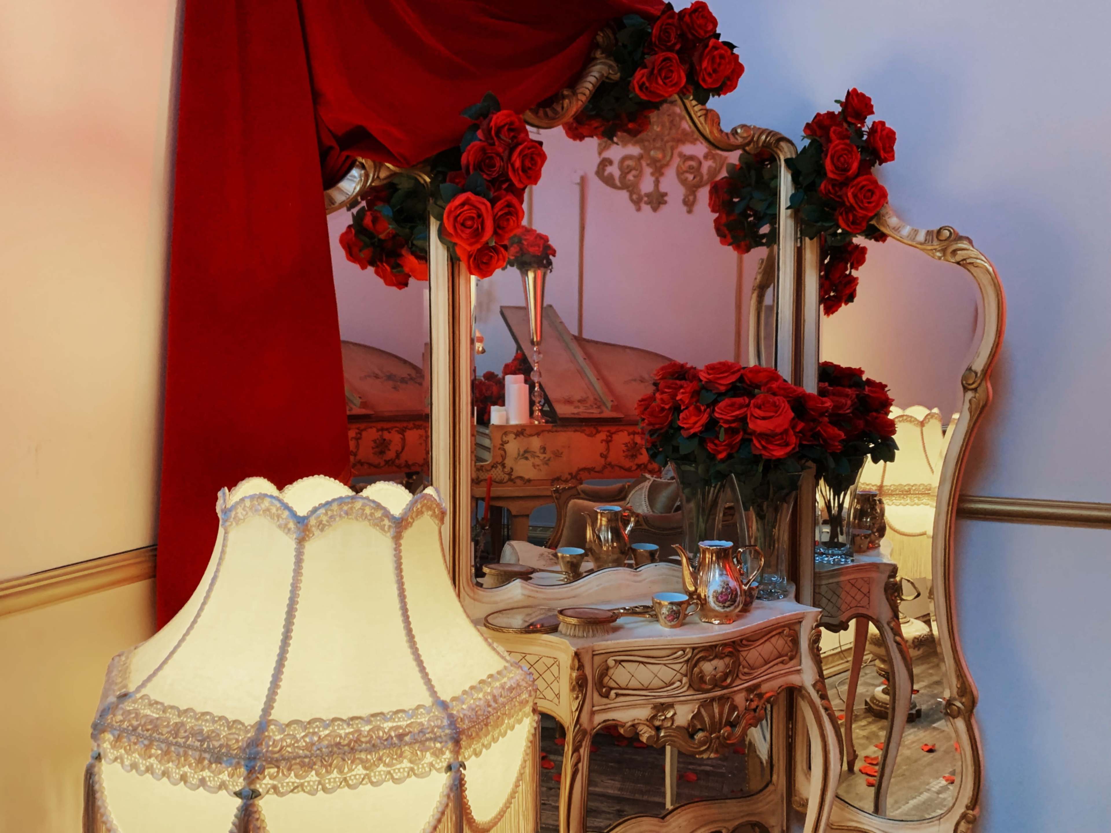 An ornate mirror surrounded by red roses and a vintage lamp, set against a backdrop of flowing red fabric.