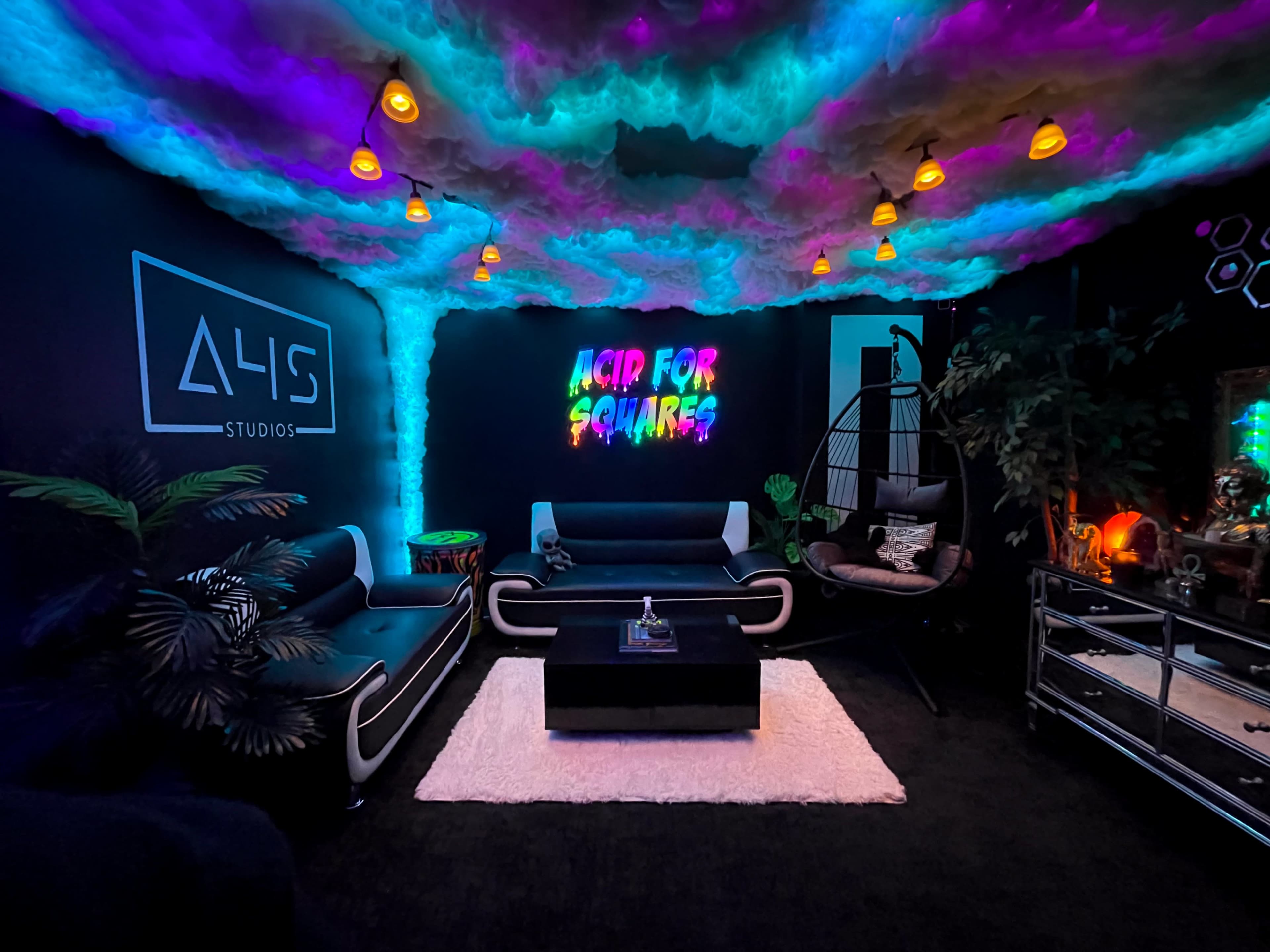 The image shows a modern lounge area with colorful lighting, featuring black sofas, a coffee table, and decorative plants under a cloud-like ceiling.