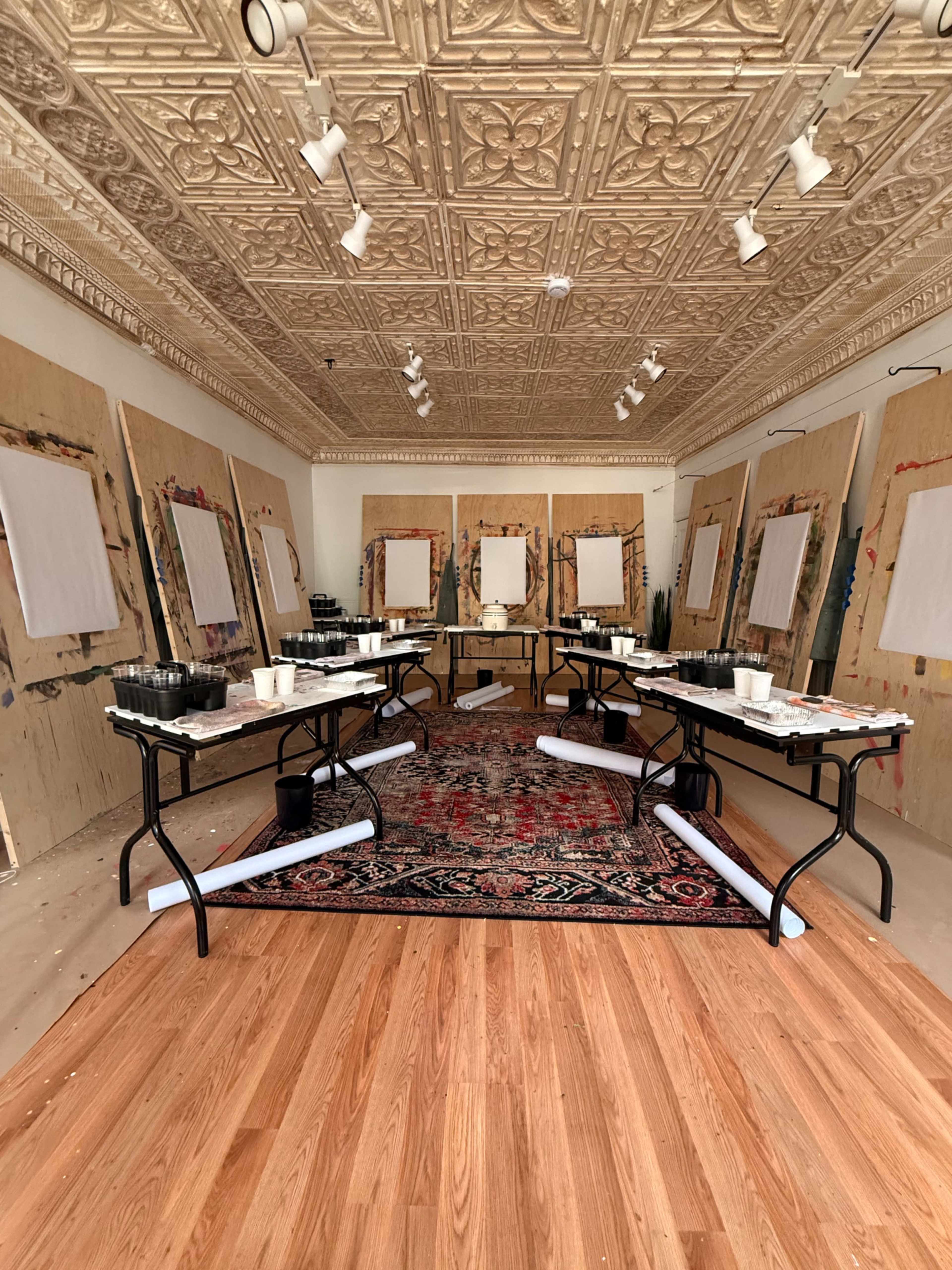 The image shows an art studio with several tables set up in a light-filled room, surrounded by blank canvases mounted on wooden boards.
