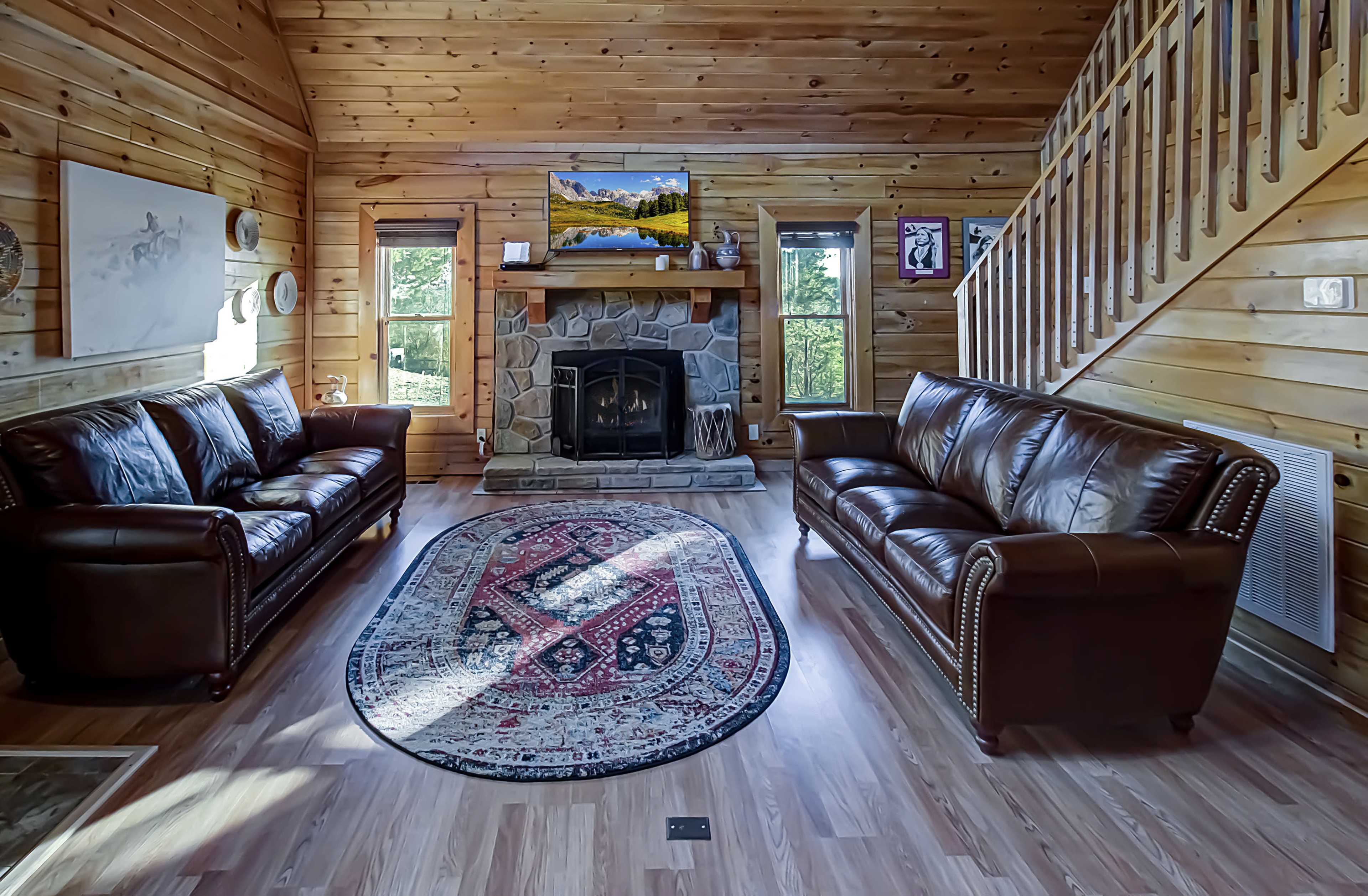 The image shows a cozy wooden living room featuring two brown leather sofas, a stone fireplace with a mantel, a patterned area rug, and a staircase in the background.