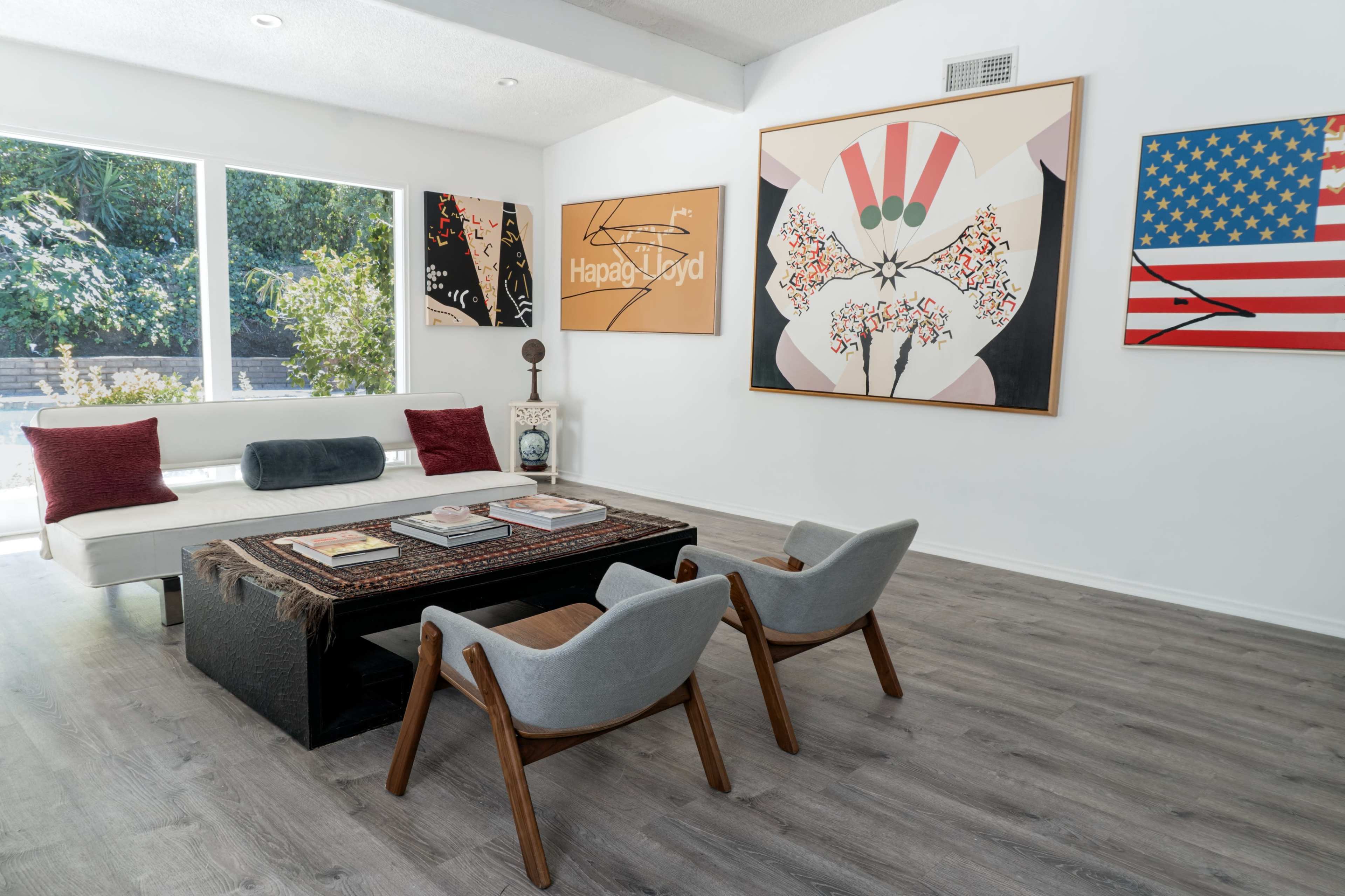 The living room features a white couch, two mid-century chairs, and a coffee table, with walls adorned by colorful artwork and large windows that provide natural light.
