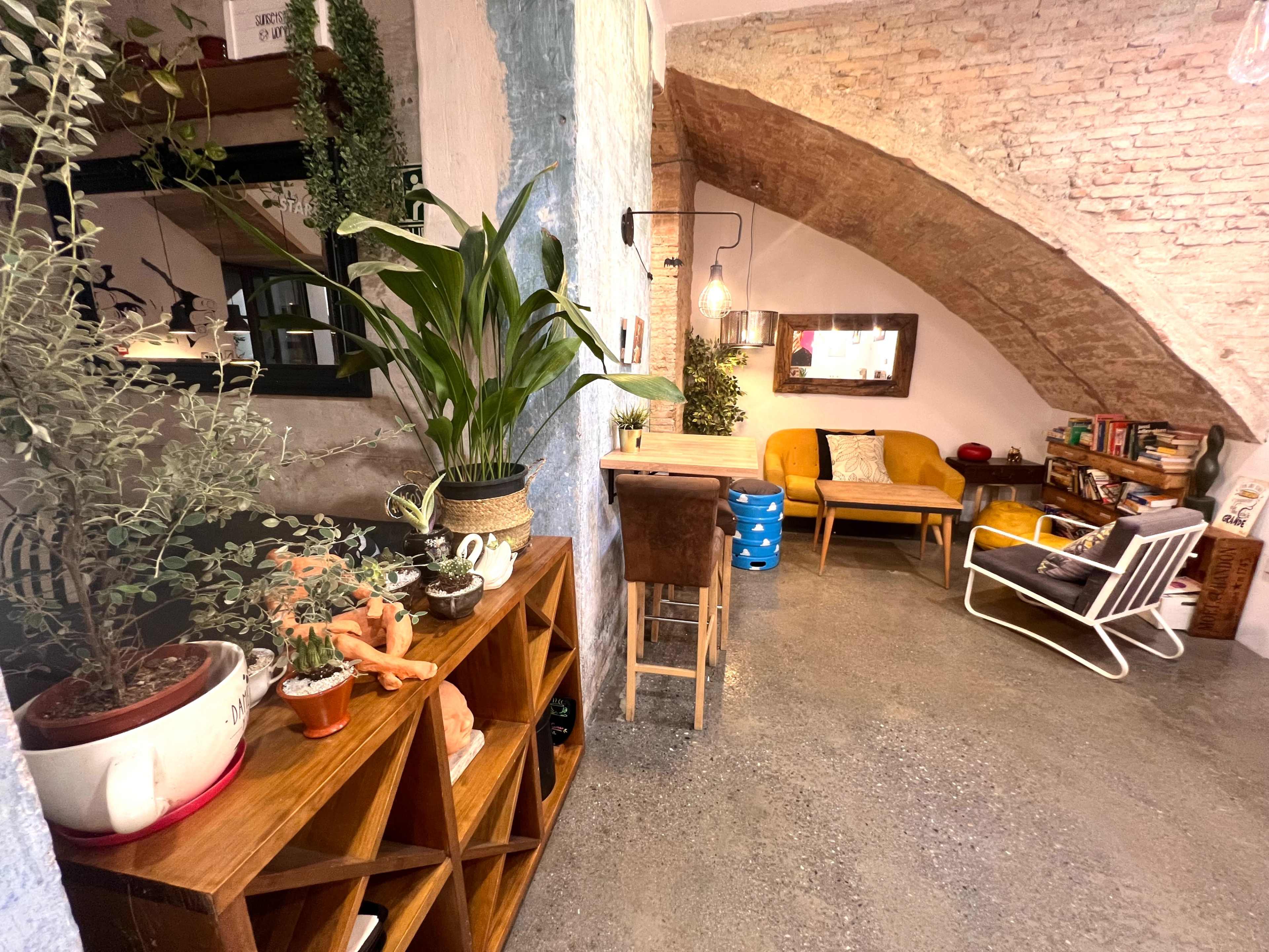 The image shows a cozy interior space featuring wooden furniture, plants, a curved brick ceiling, and a seating area with a yellow couch and a rocking chair.