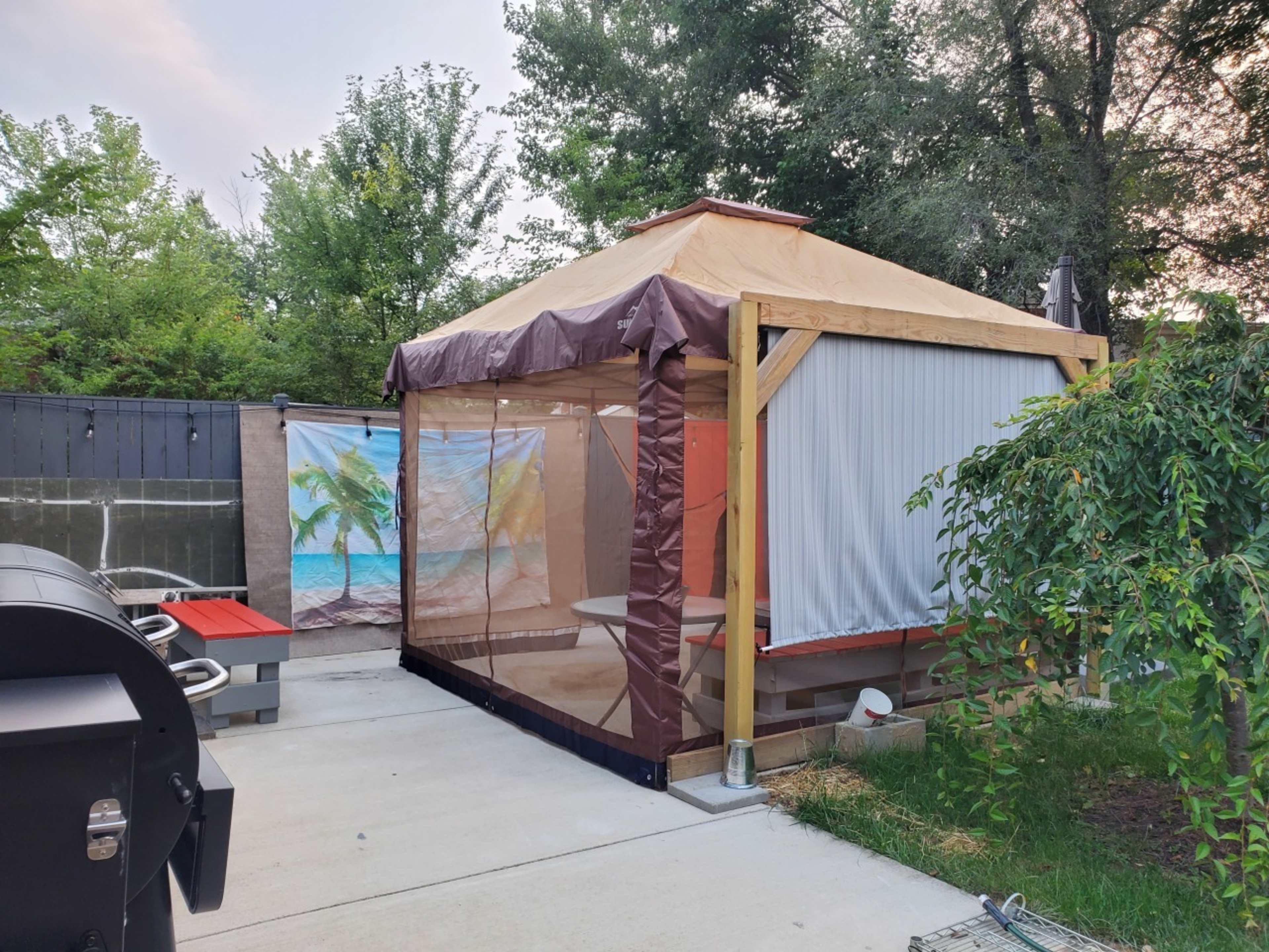 A screened gazebo with a tan roof and brown trim is situated on a concrete patio surrounded by greenery and outdoor furniture.