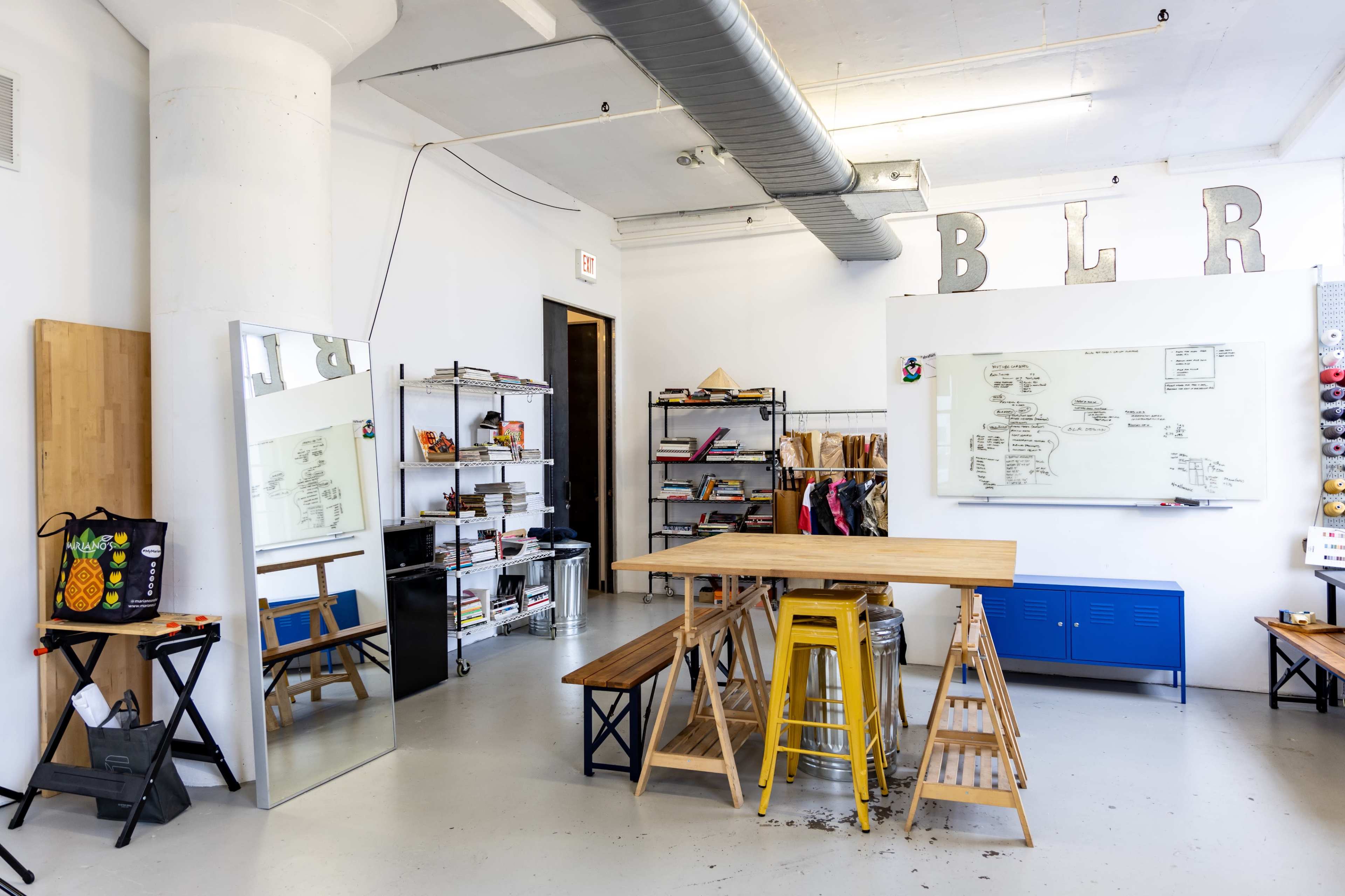 A bright, spacious room features a large table made of wooden planks, metal stools, a mirror, shelves filled with books and materials, and a whiteboard on the wall.