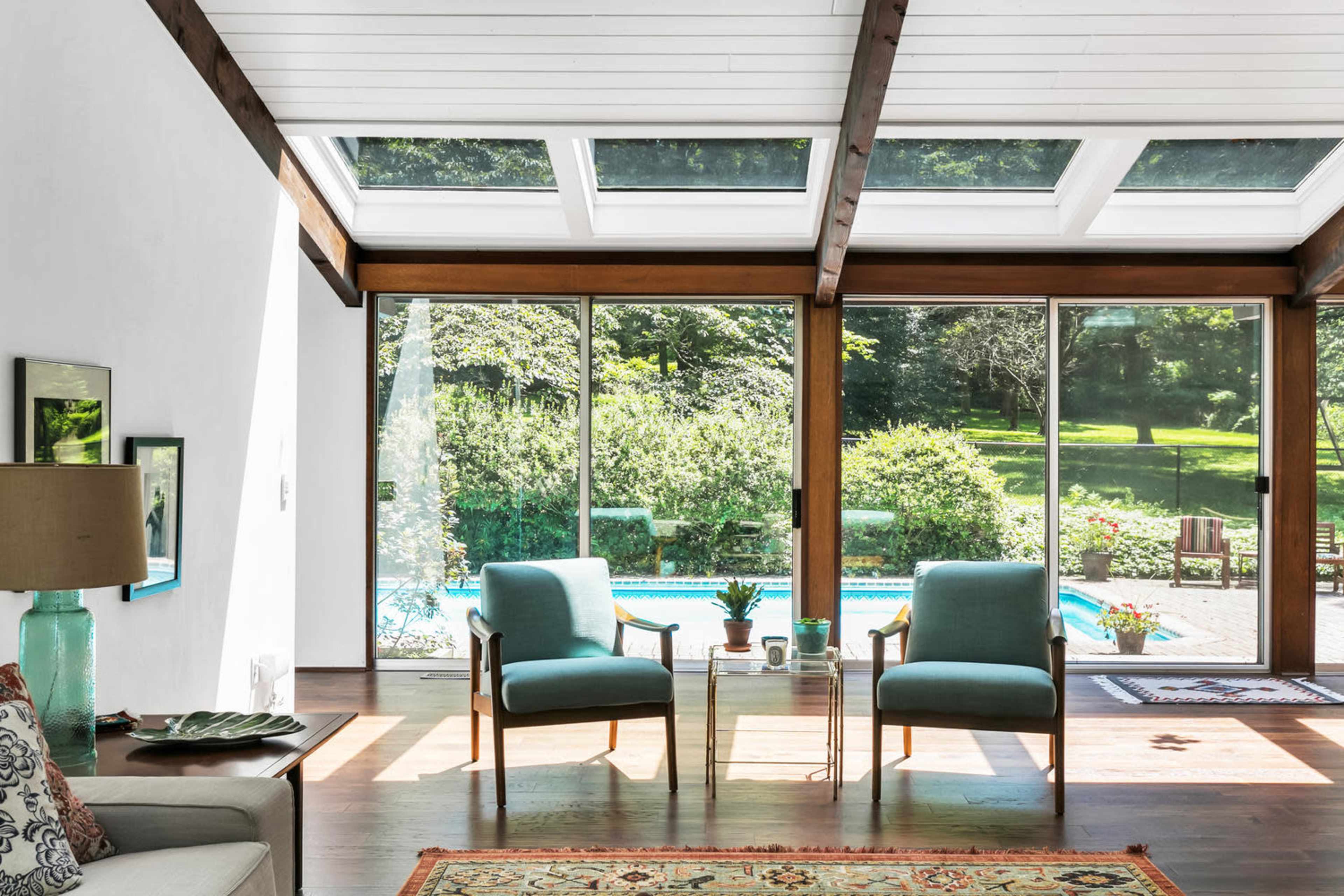 A sunlit room features two chairs, a small table, and large windows overlooking a pool and lush greenery outside.