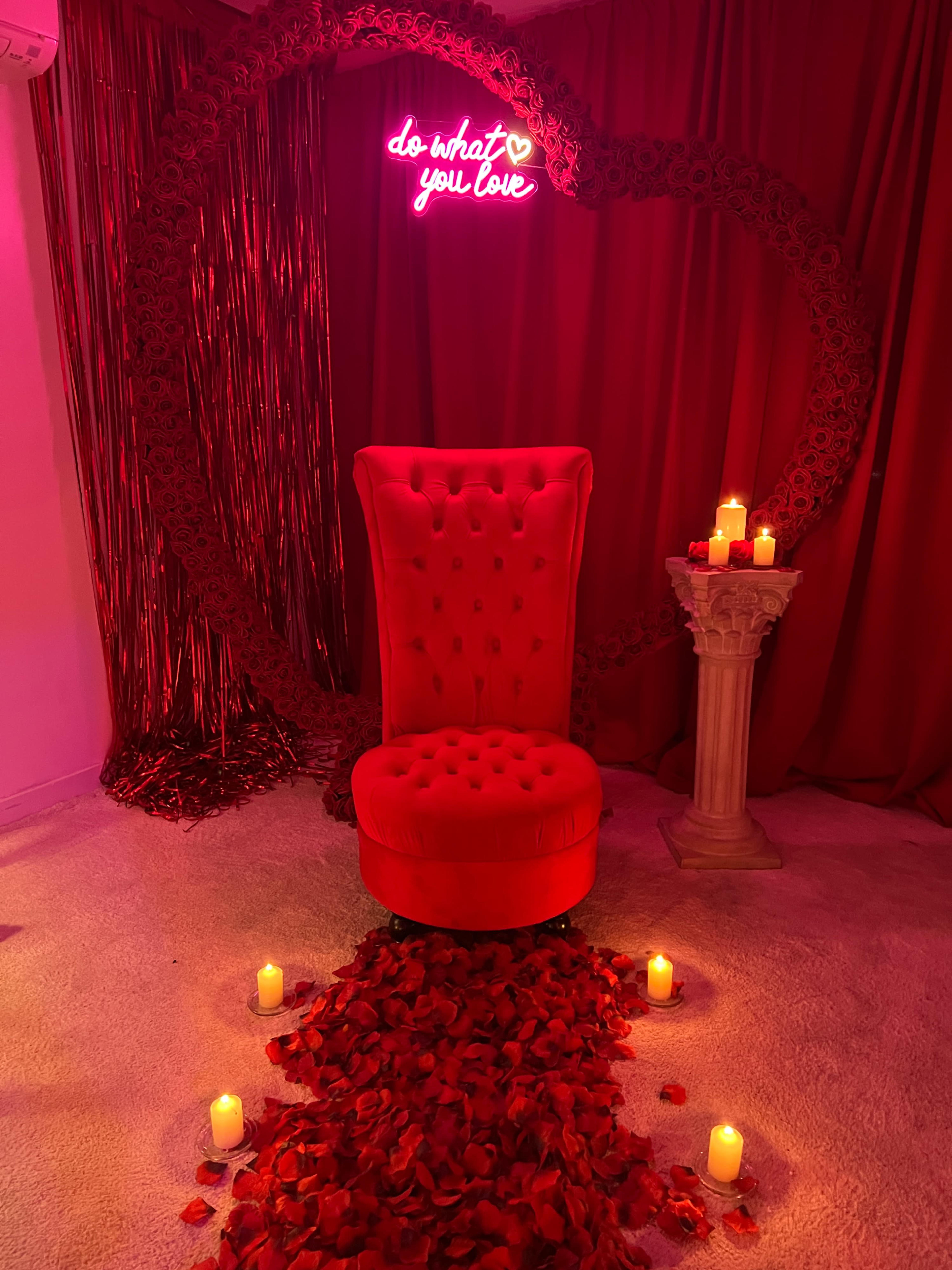 A red velvet chair positioned in front of a heart-shaped arrangement of roses and candles, with a neon sign that reads "do what you love."