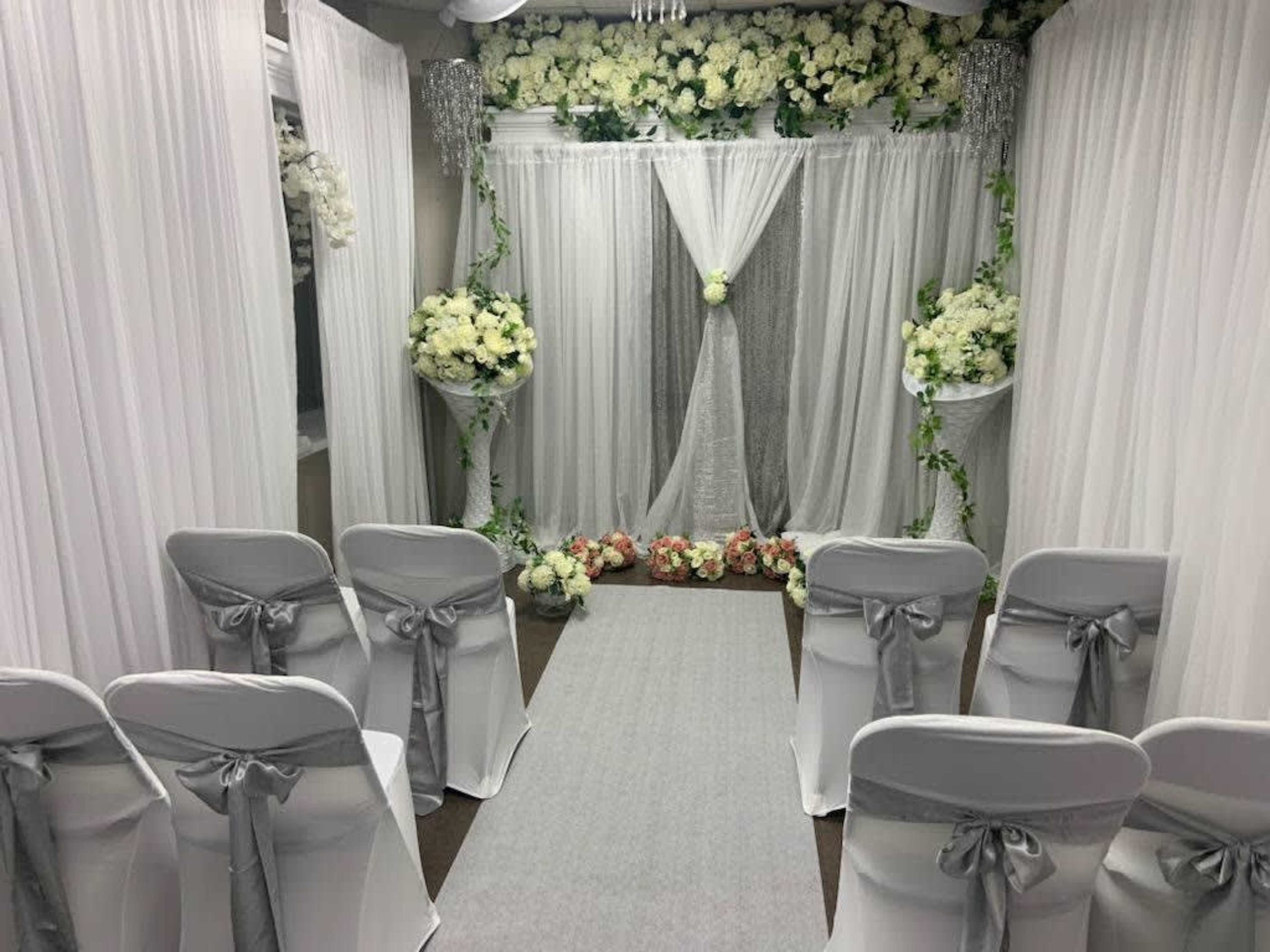 The image shows a wedding ceremony setup with white drapes, floral arrangements, and chairs arranged along a gray aisle.