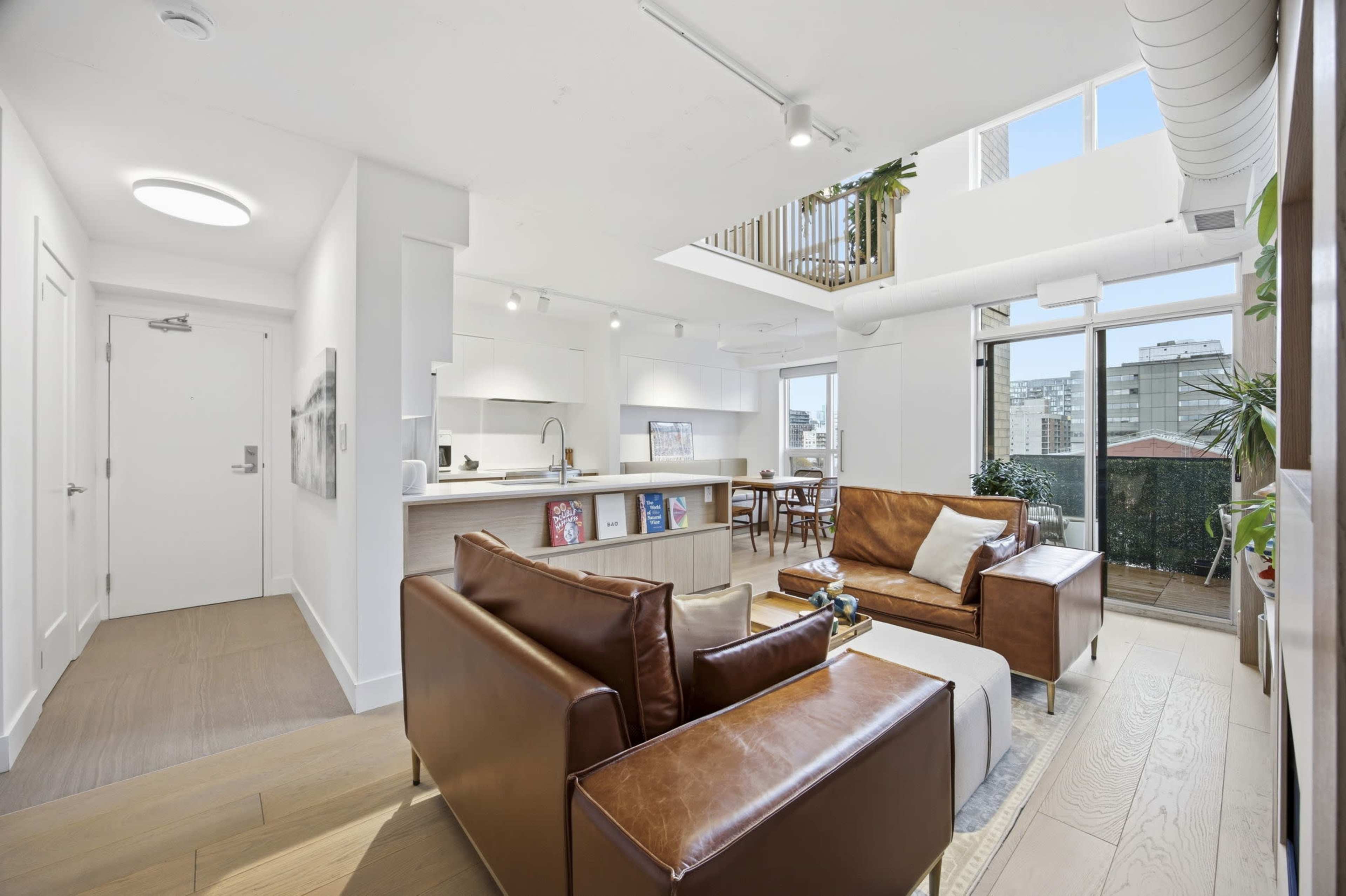 The image shows a modern living room with brown leather sofas, a wooden dining area, and large windows offering a view of the city.