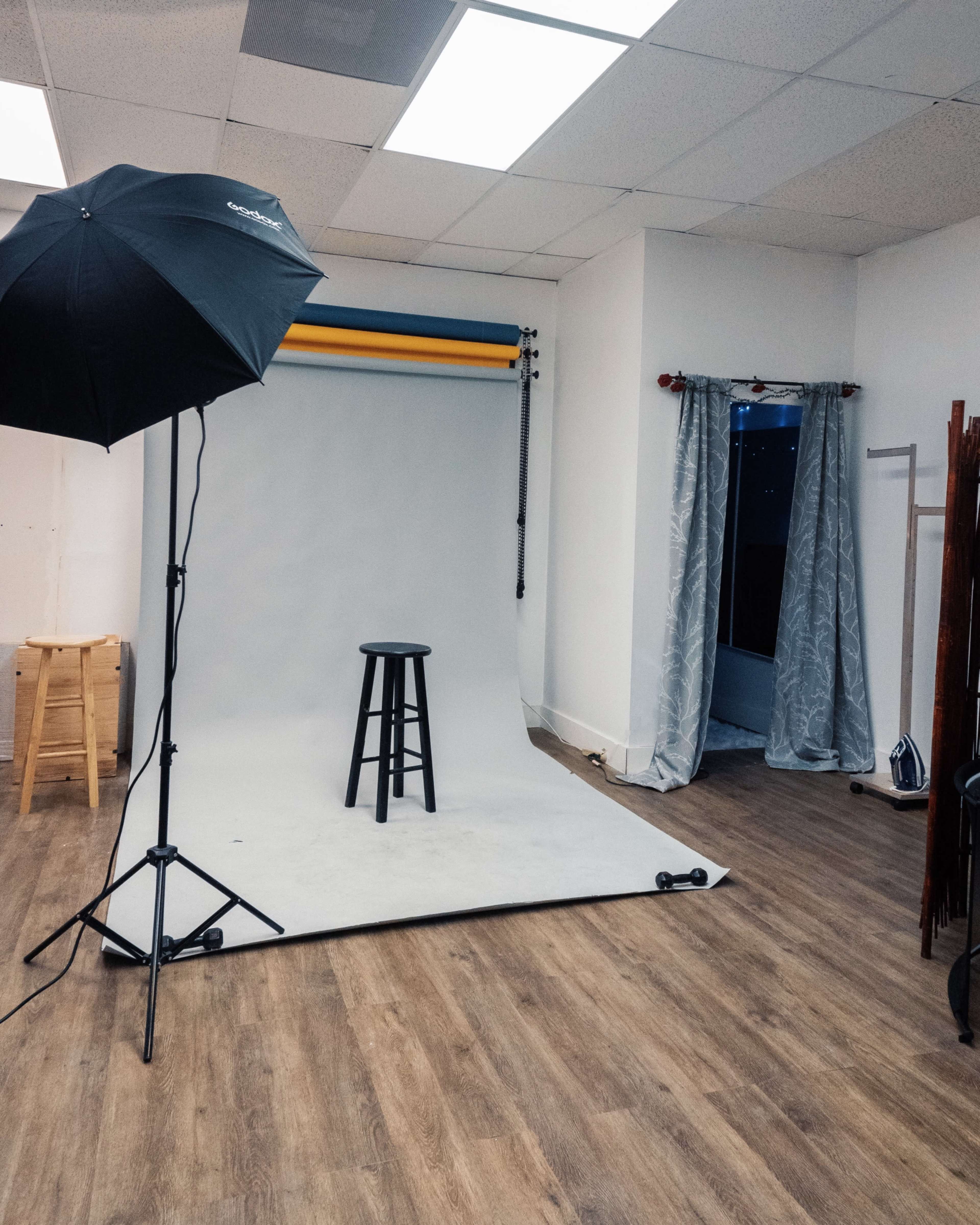 A photography studio features a gray backdrop, a black stool in the center, and an umbrella light stand positioned to the side.
