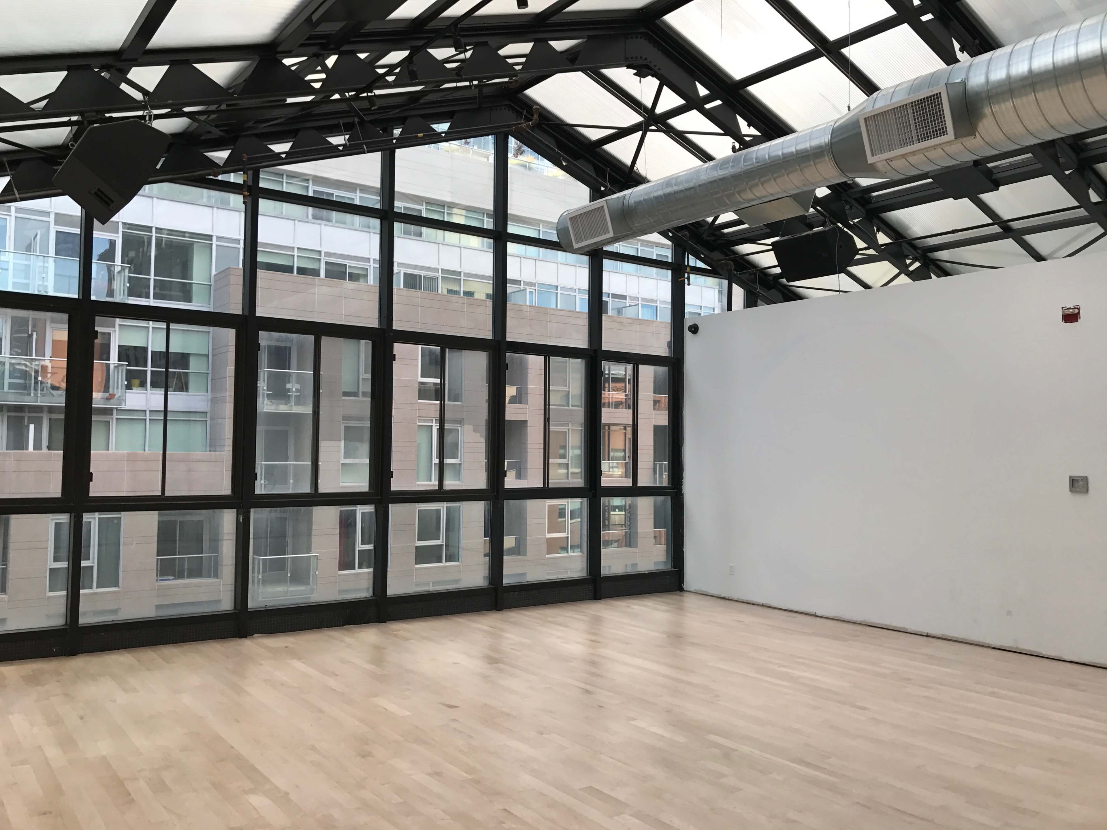 The image shows an empty room with wood flooring and large windows showcasing a view of adjacent buildings.