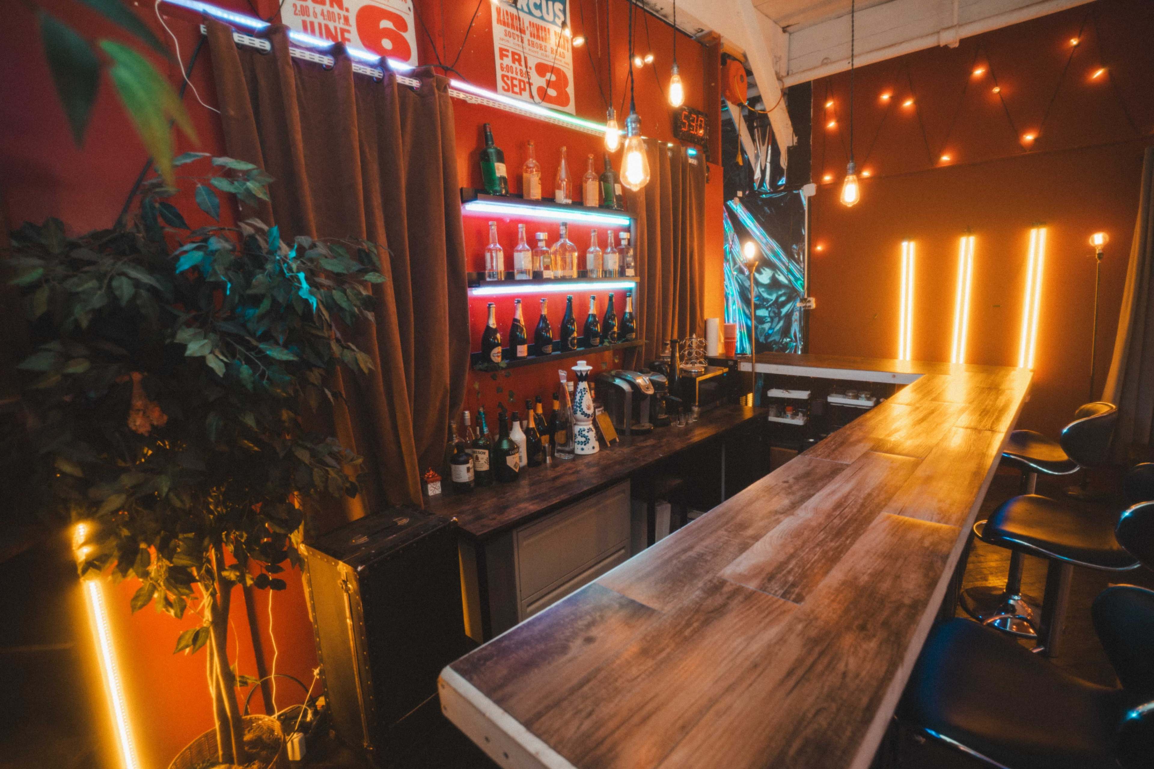 The image shows a well-lit bar area featuring a wooden countertop, various bottles on shelves, and decorative lighting.