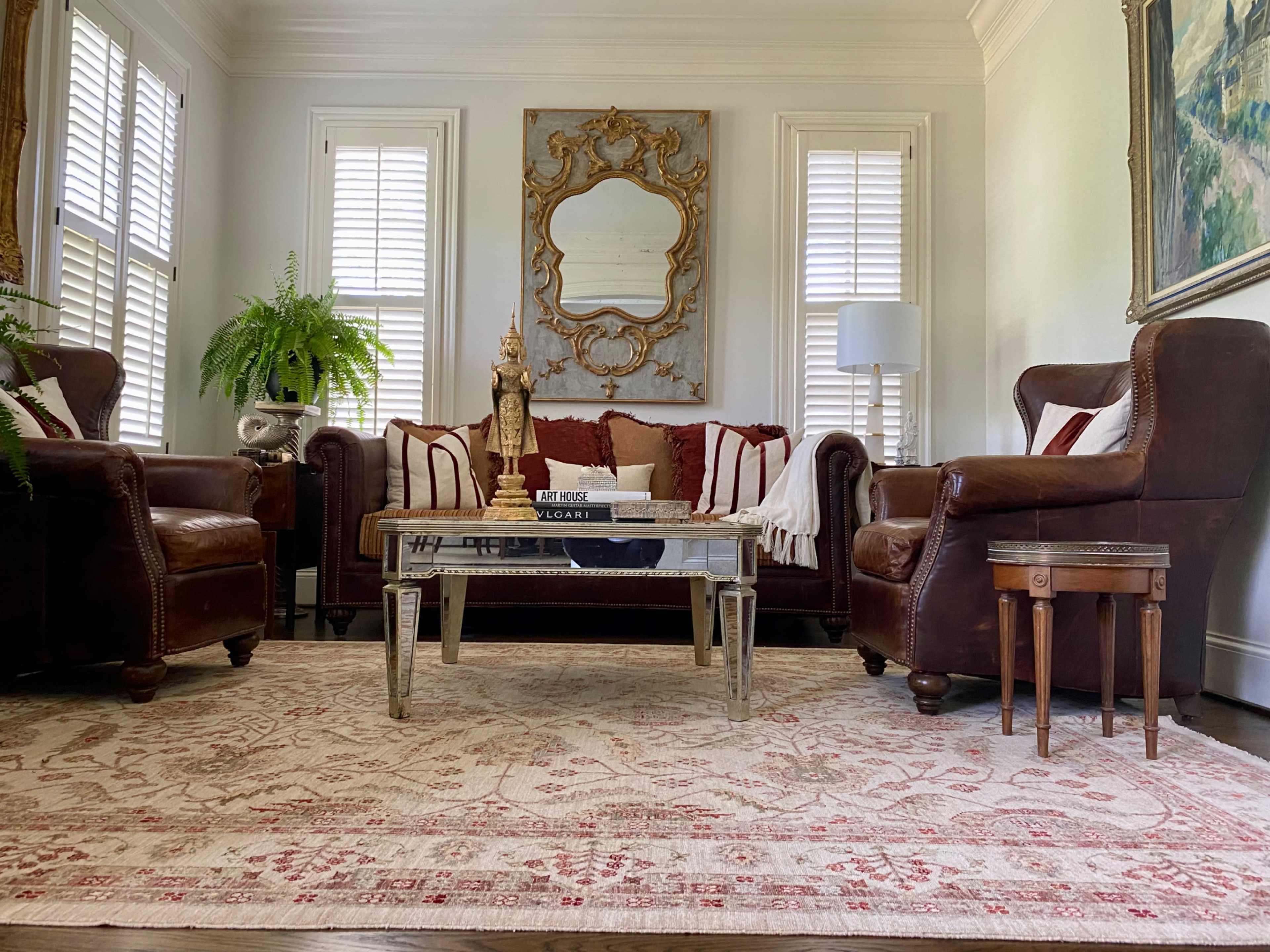 A cozy living room features a brown leather sofa with white and red accent pillows, flanked by two matching armchairs, a glass coffee table, and a large mirror above, with light streaming through the window shutters.