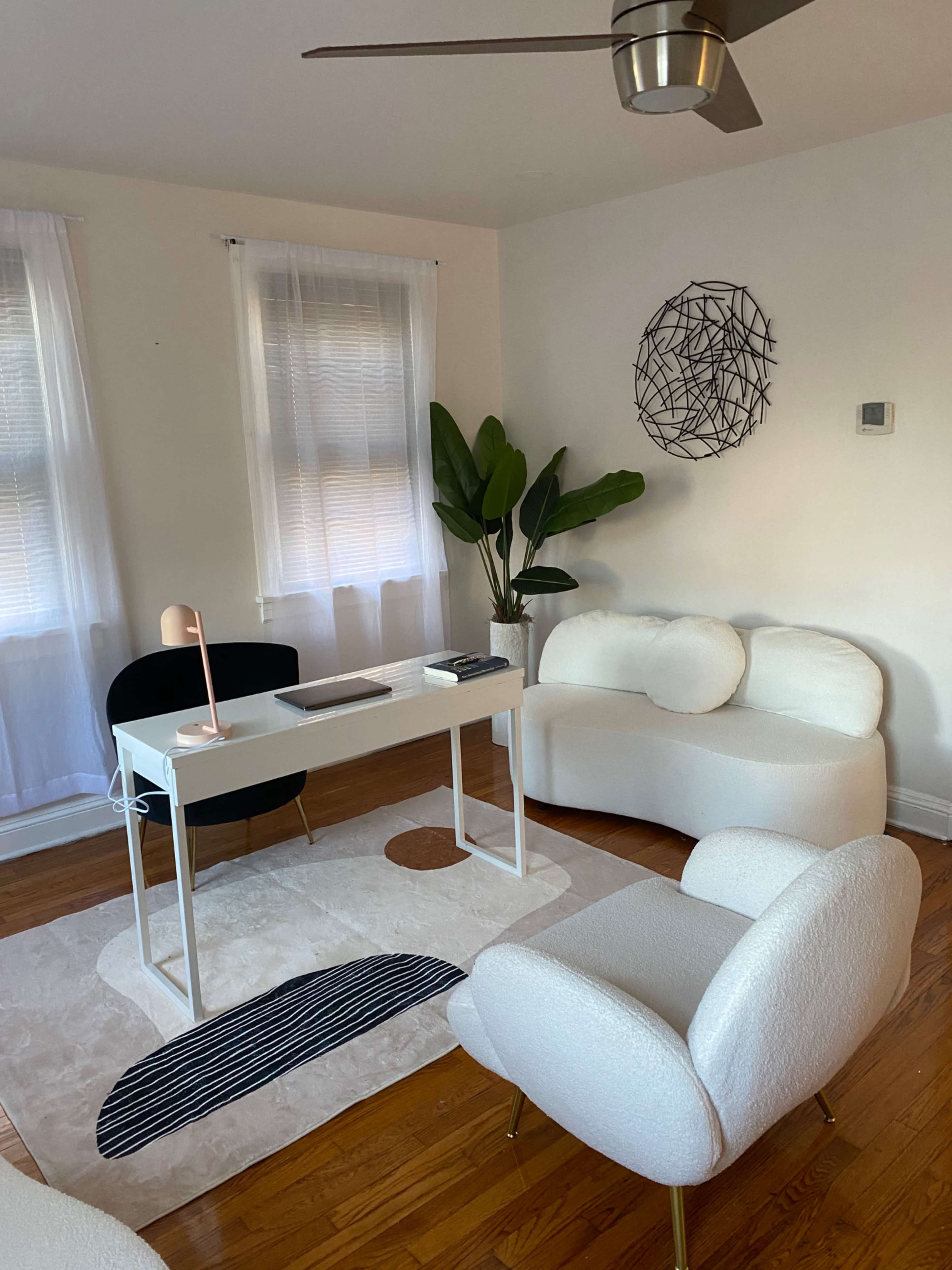 A bright, minimalist office with white furniture, a desk facing a large window, and a potted plant in the corner.