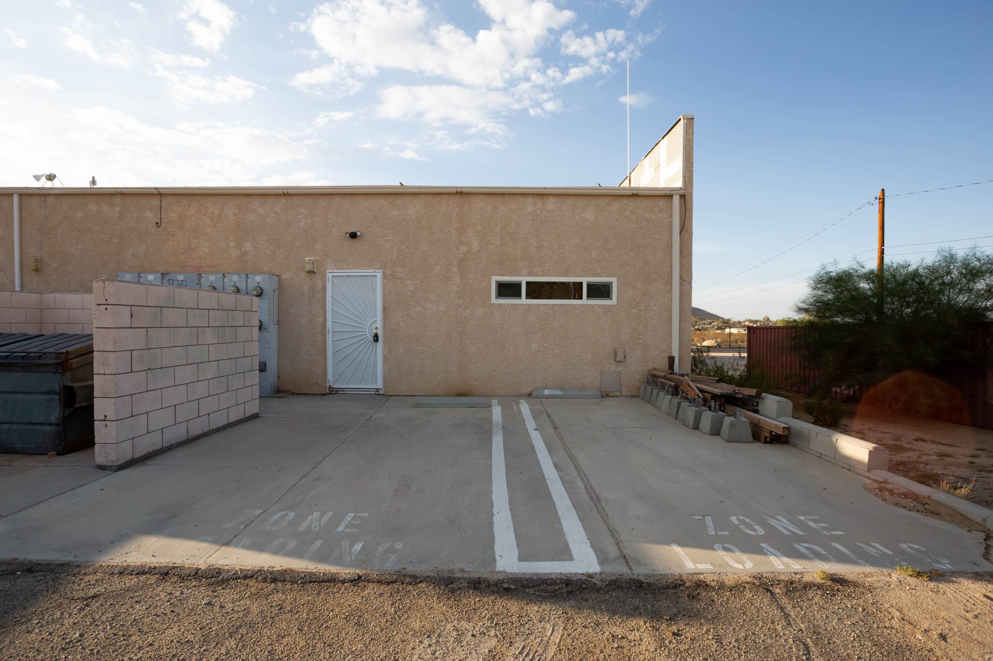 A beige building with a single door and a window, flanked by a loading zone marked on the concrete pavement.