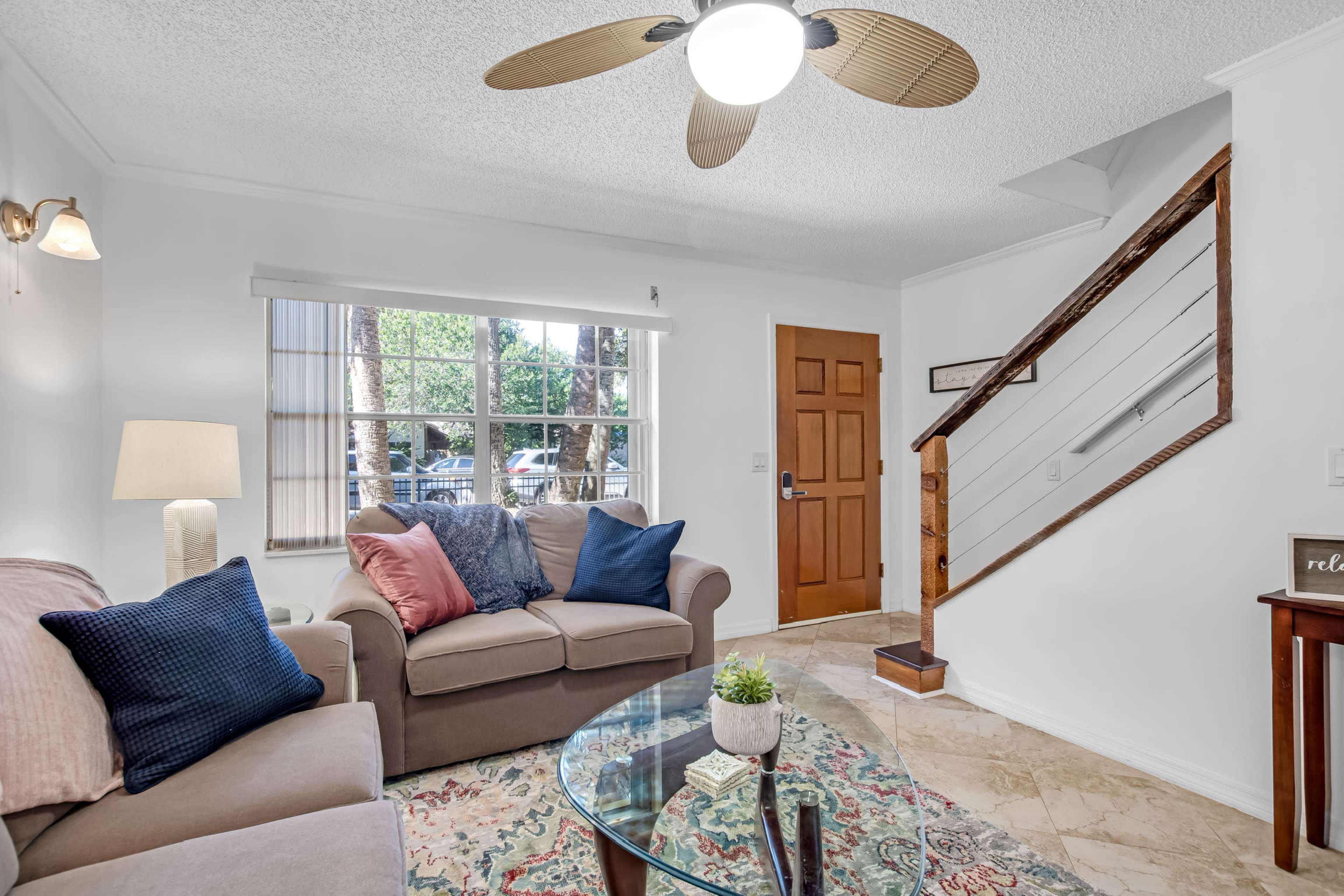 A living room features a sofa, a coffee table with a plant, a ceiling fan, and a staircase leading to an upper level.