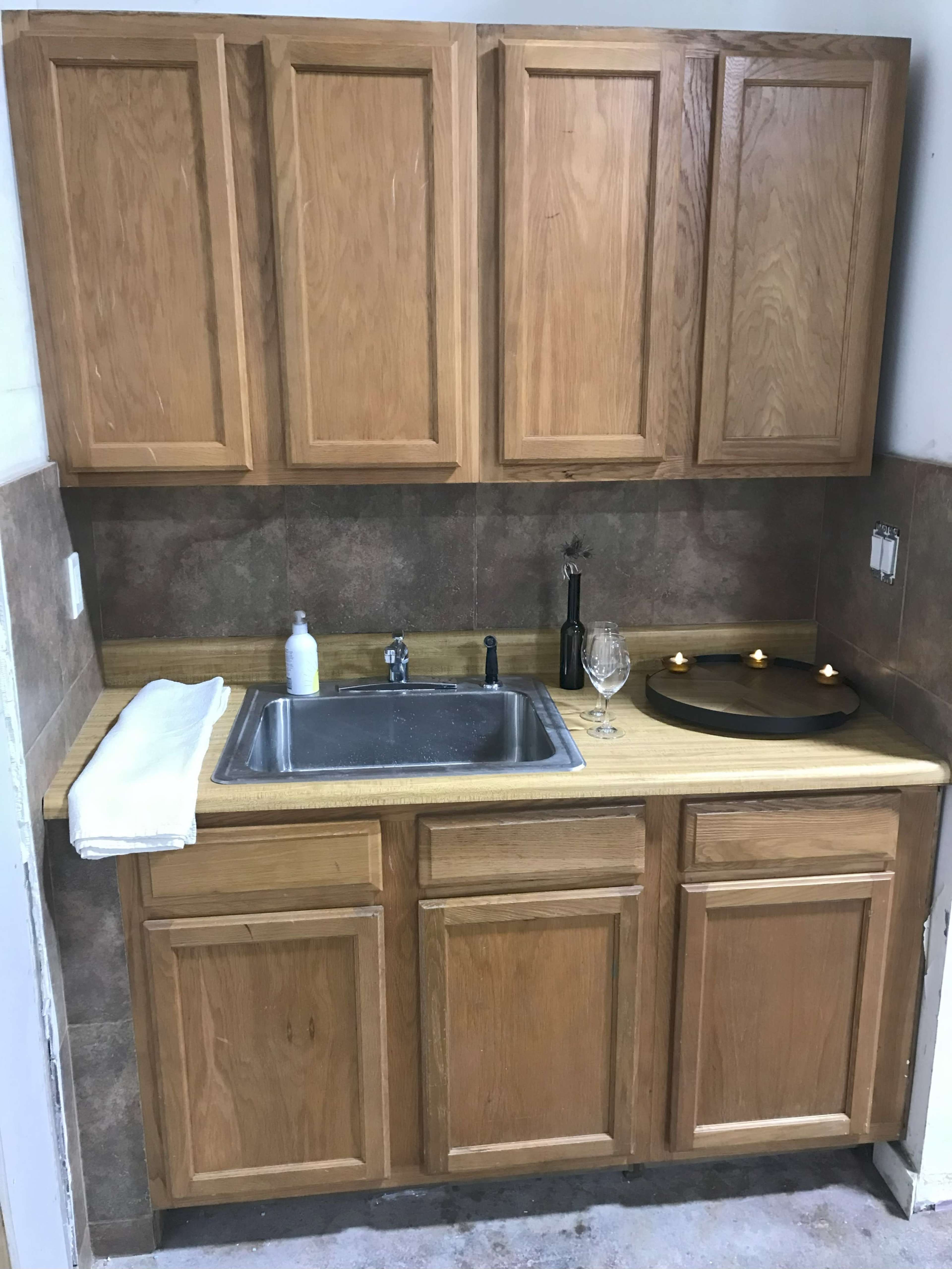 The image shows a small kitchen area with wooden cabinets above a stainless steel sink, a countertop, a bottle, a glass, and three candles.