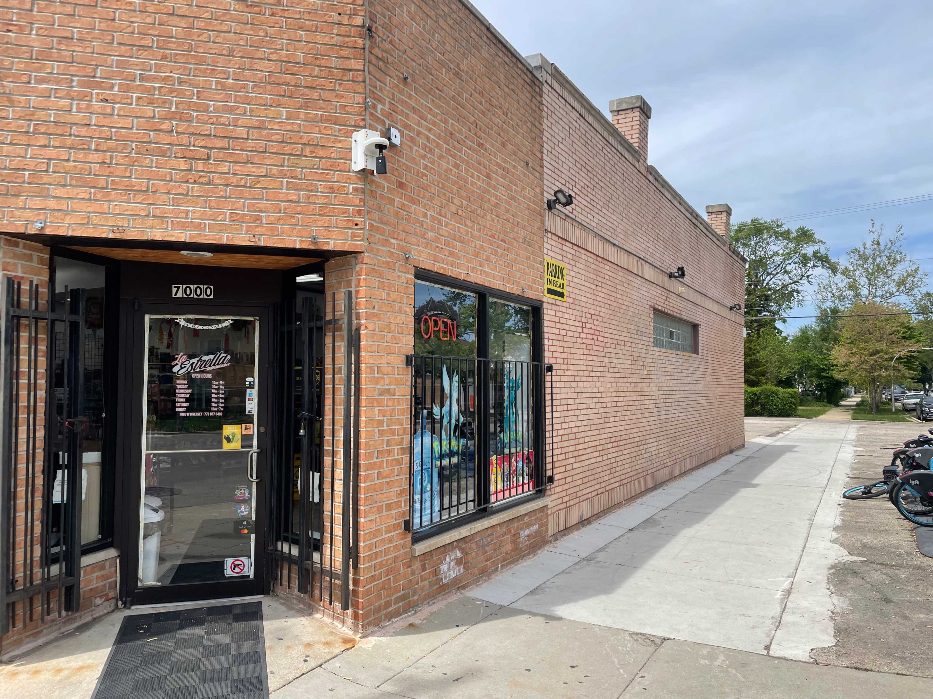 A brick storefront with an open sign in the window and a sidewalk running alongside it.