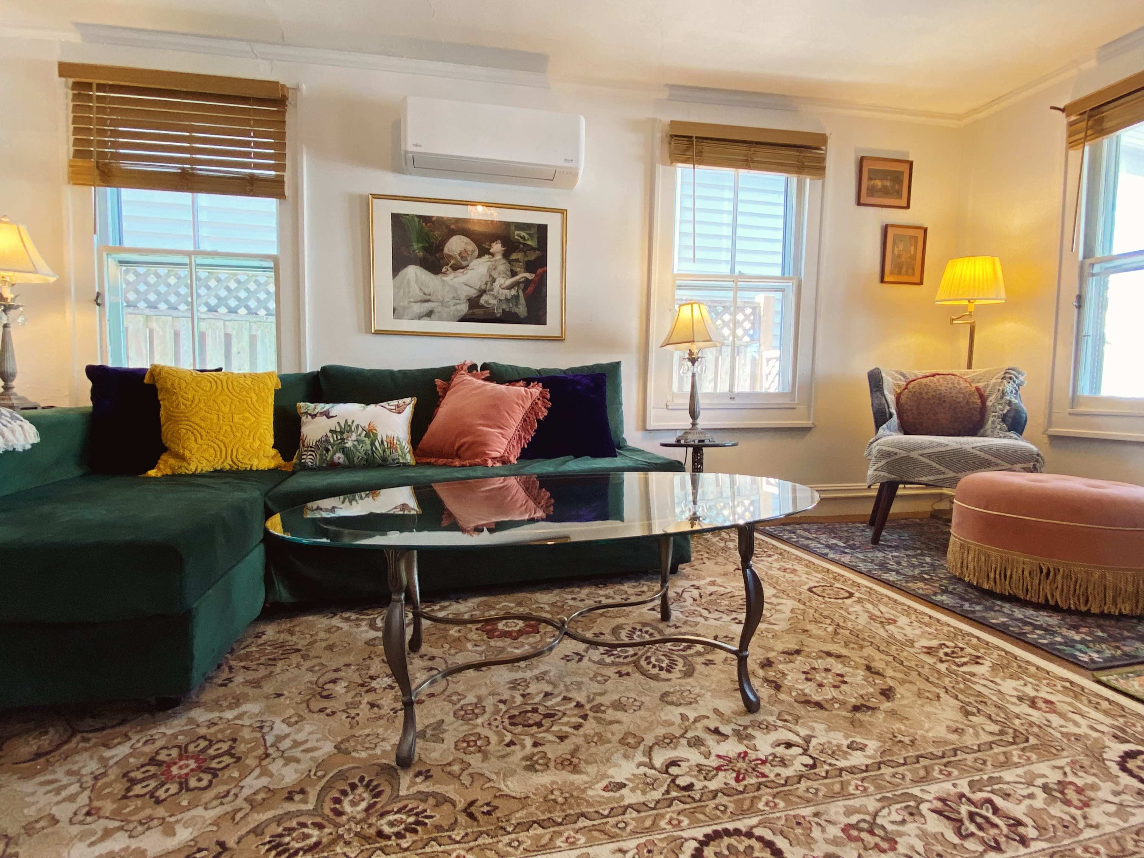 A cozy living room featuring a green sectional sofa adorned with colorful pillows, a glass coffee table, and a patterned rug, with ample natural light coming through two windows.