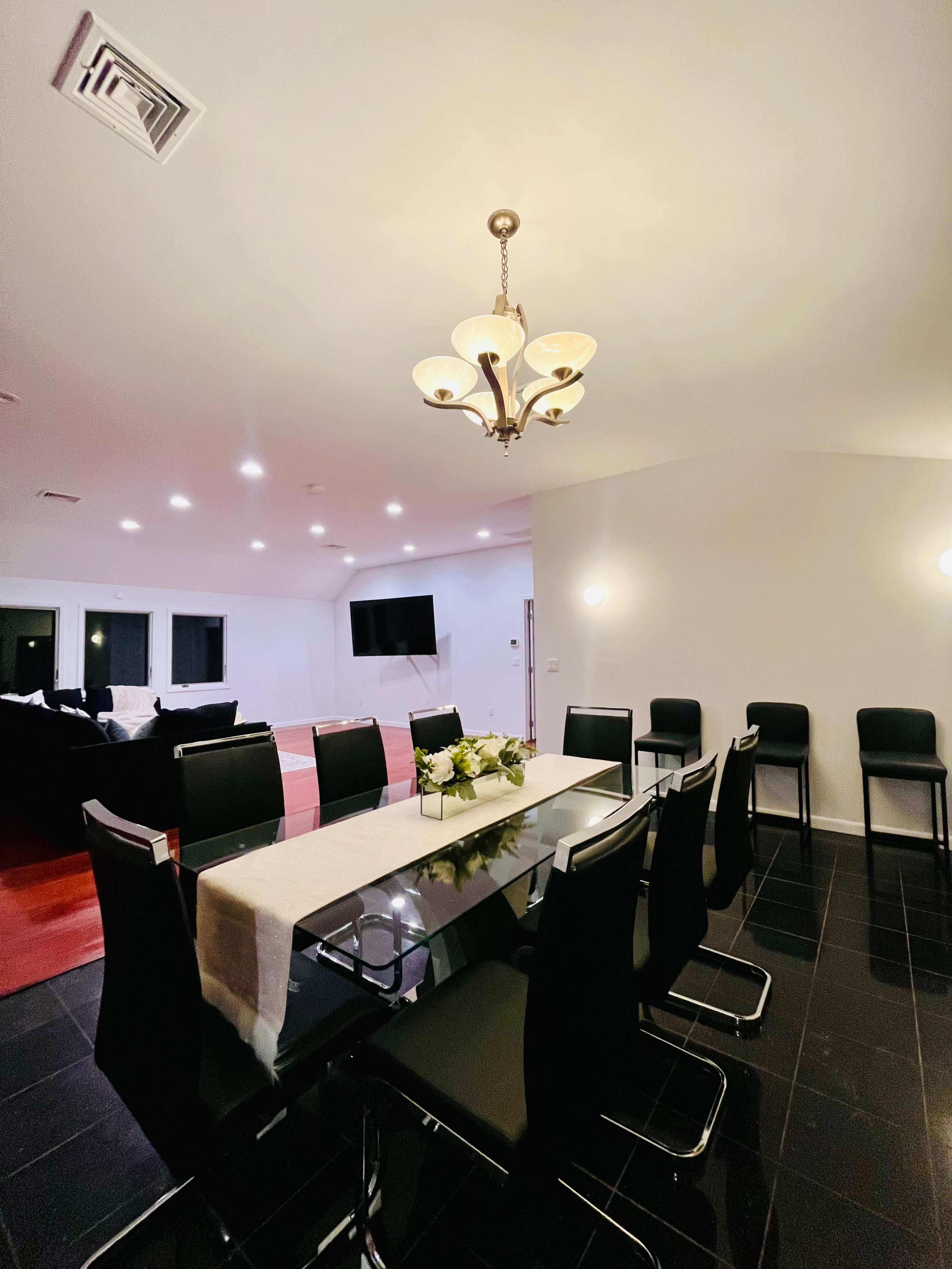 A modern dining room features a long glass table surrounded by black chairs, with a chandelier overhead and a living area in the background.