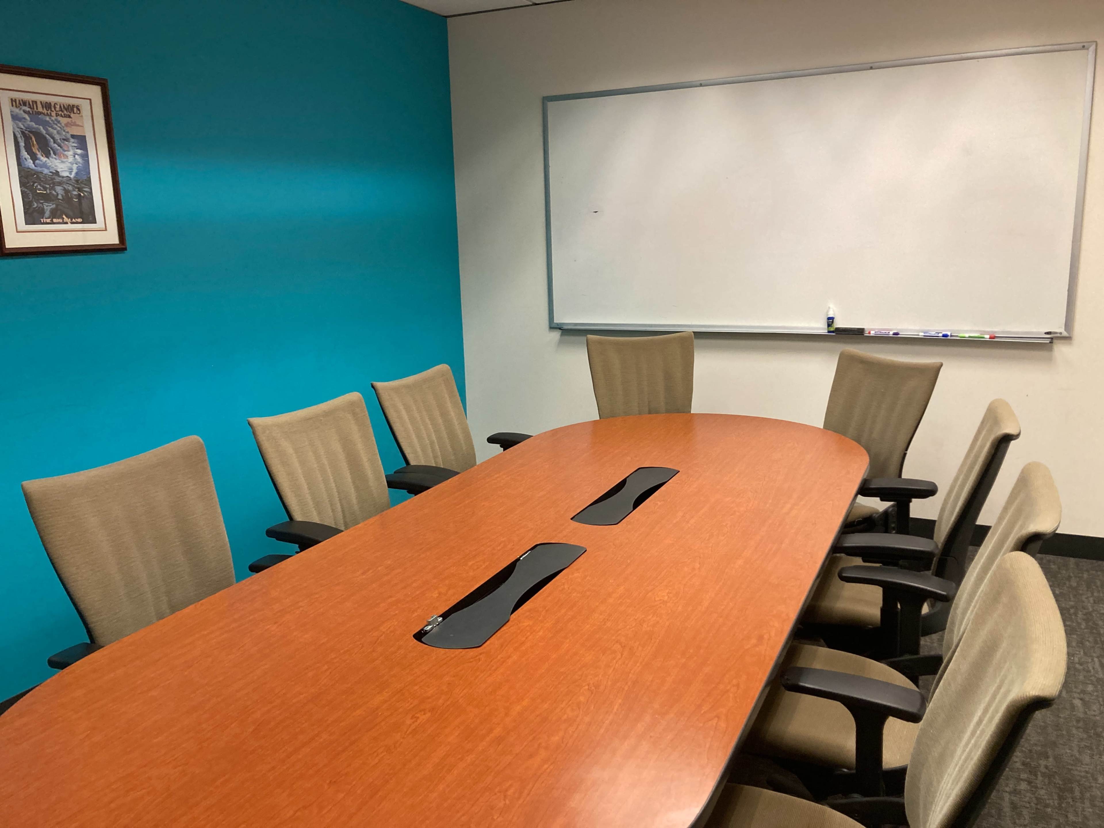The image shows a conference room with a long wooden table surrounded by beige chairs, against a turquoise wall and a whiteboard on one side.