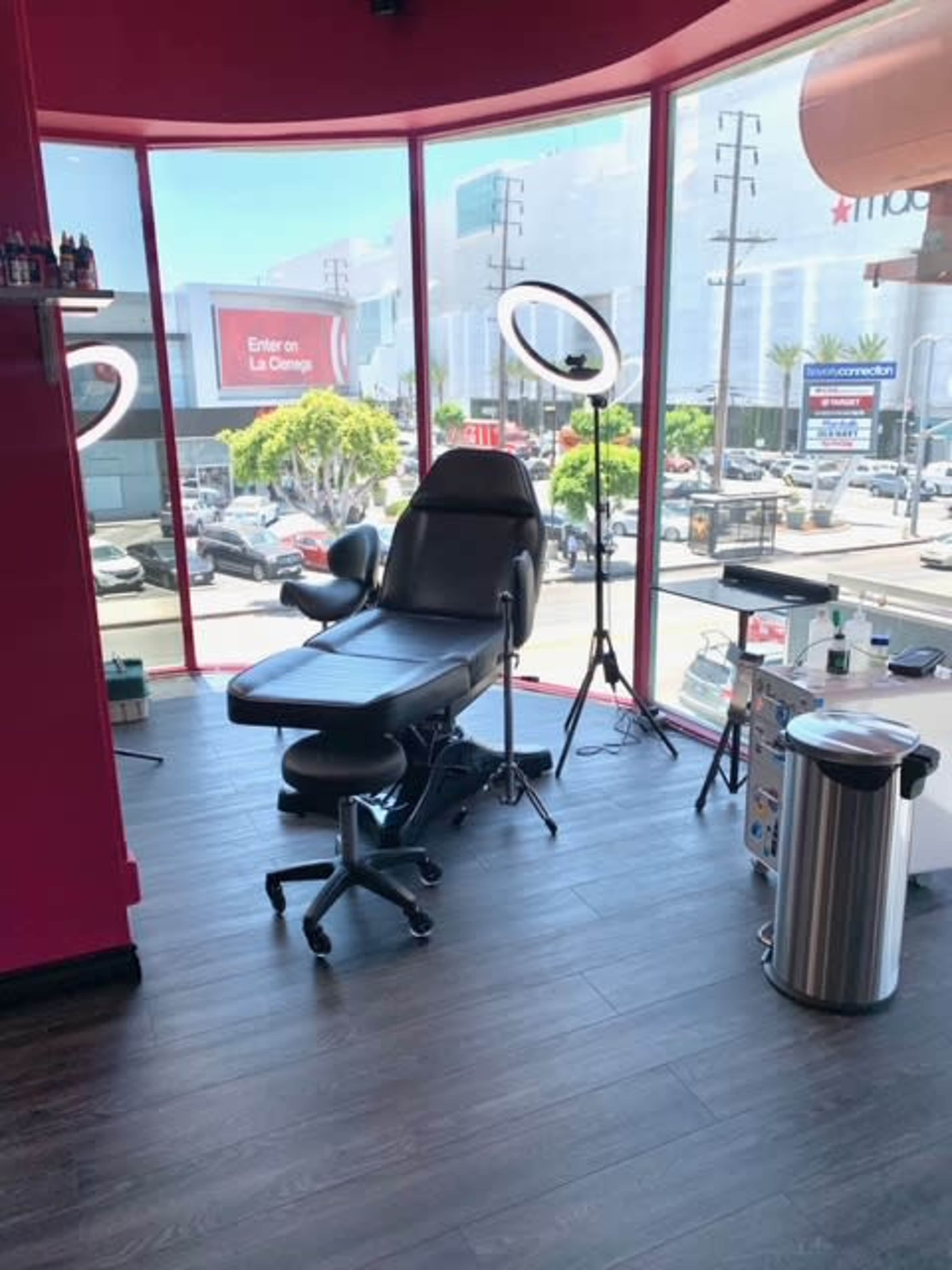 The image shows a tattoo parlor with a black tattoo chair positioned near a large window, a ring light beside it, and a street view outside.
