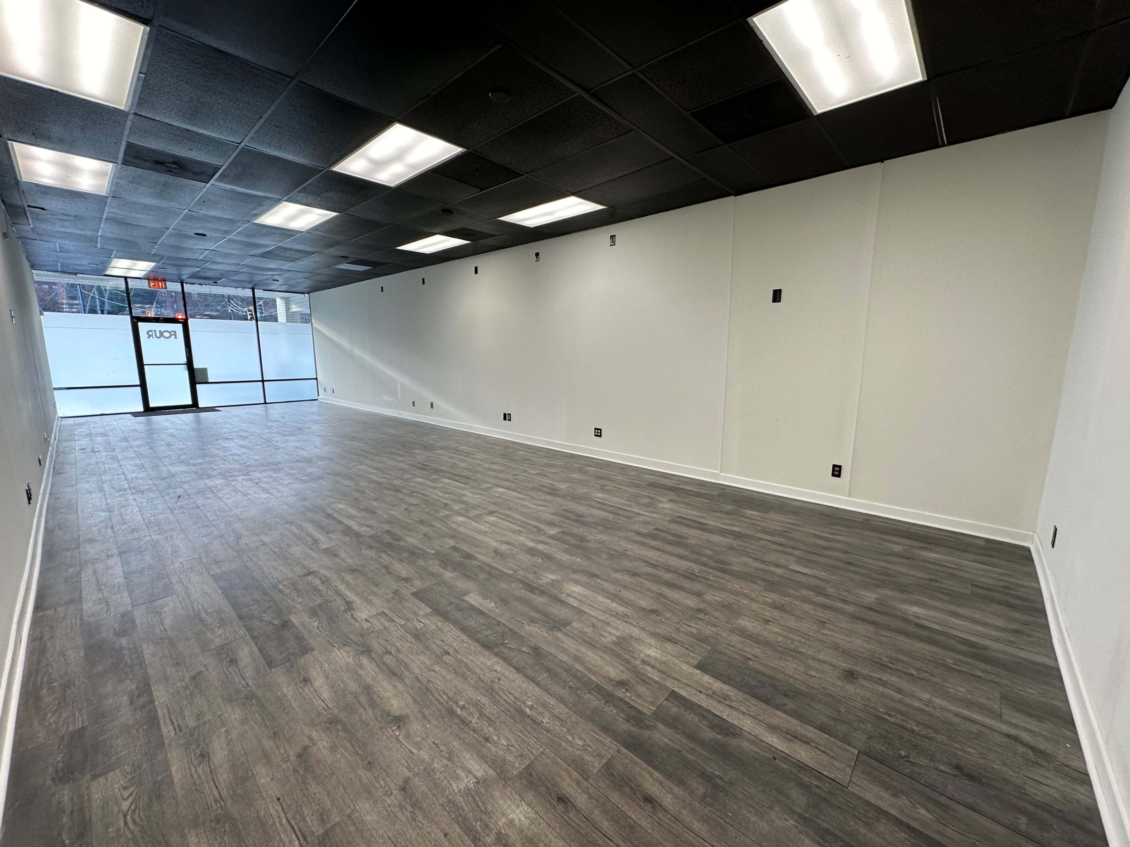 An empty interior space with gray laminate flooring and white walls, featuring a large front window and a door labeled "EXIT."