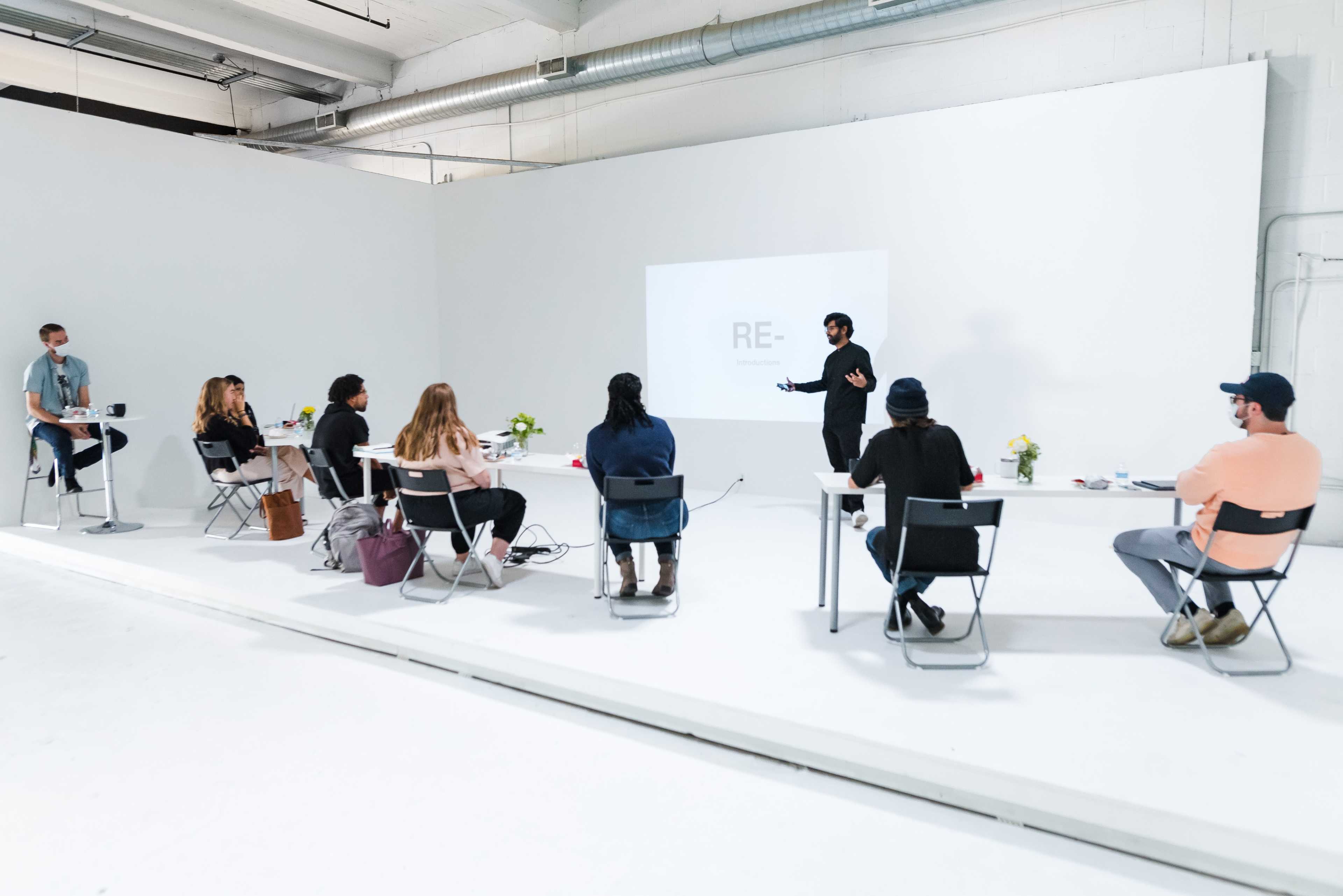 A group of people is seated at tables in a minimalist room, where a presenter is speaking in front of a projection screen.